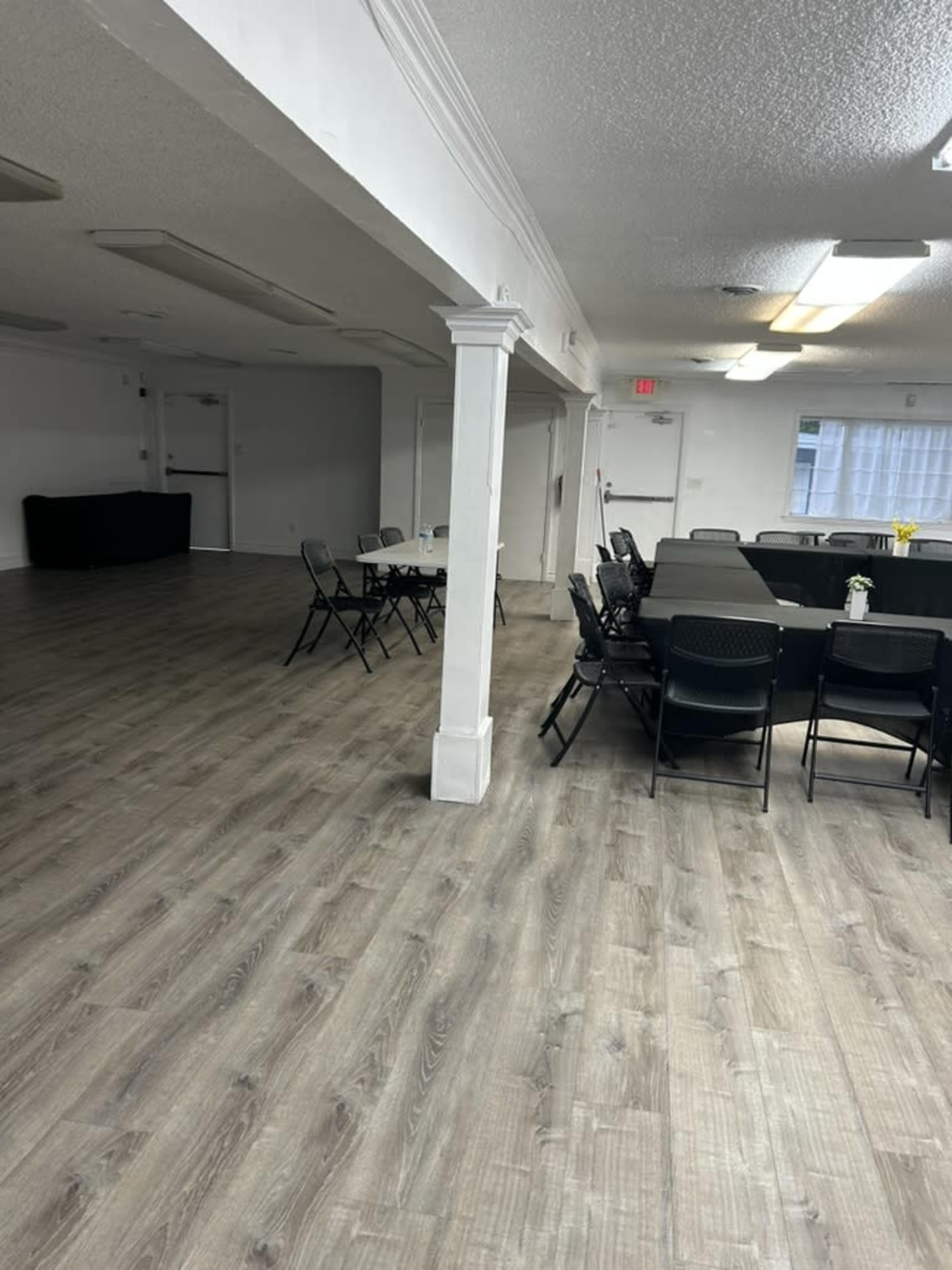 The image shows a spacious room with wooden flooring featuring several tables set with chairs, and a partitioned area in the background.