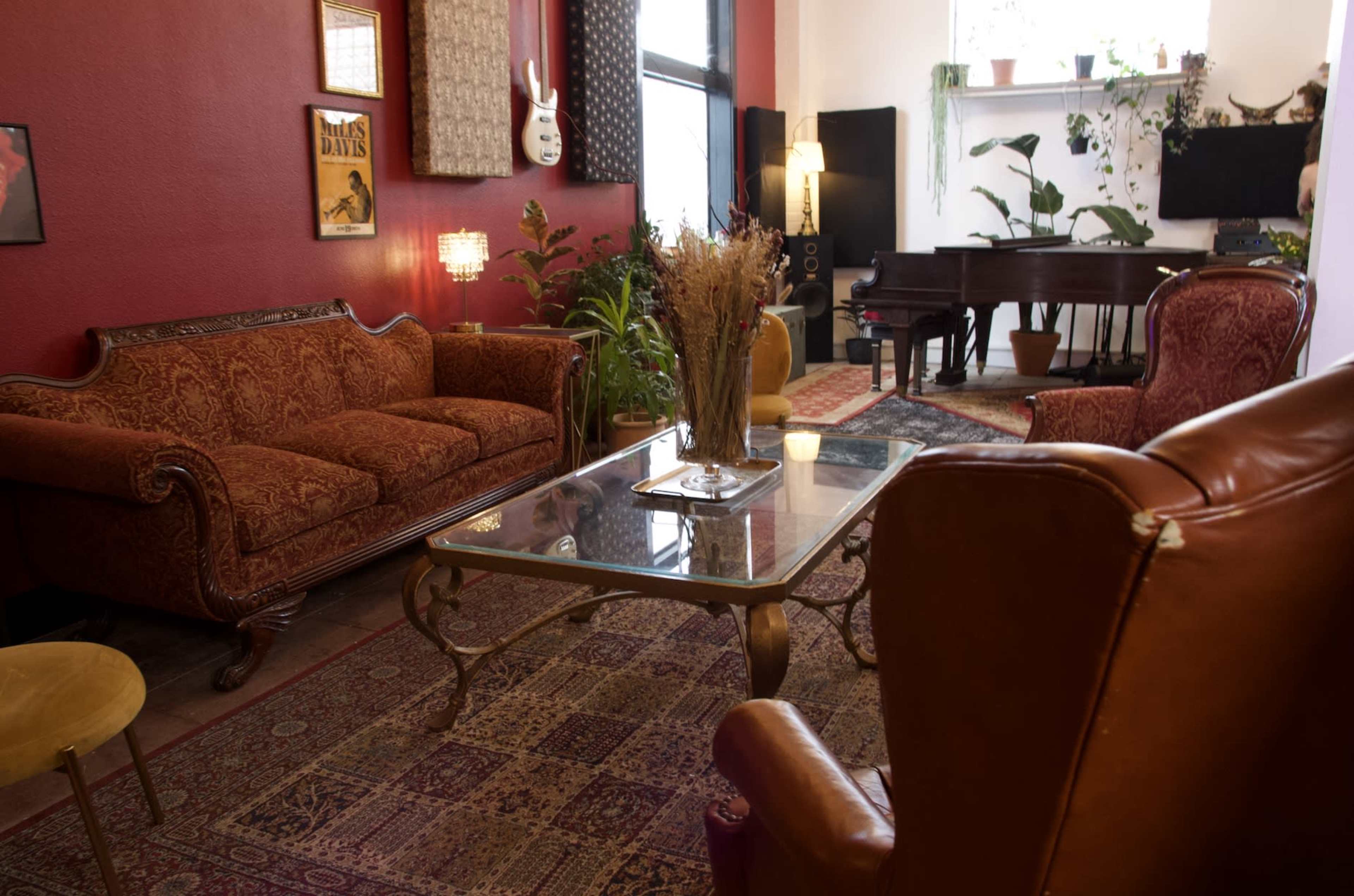 A cozy living room with ornate furniture, a glass coffee table, and a grand piano in the background, surrounded by various plants.
