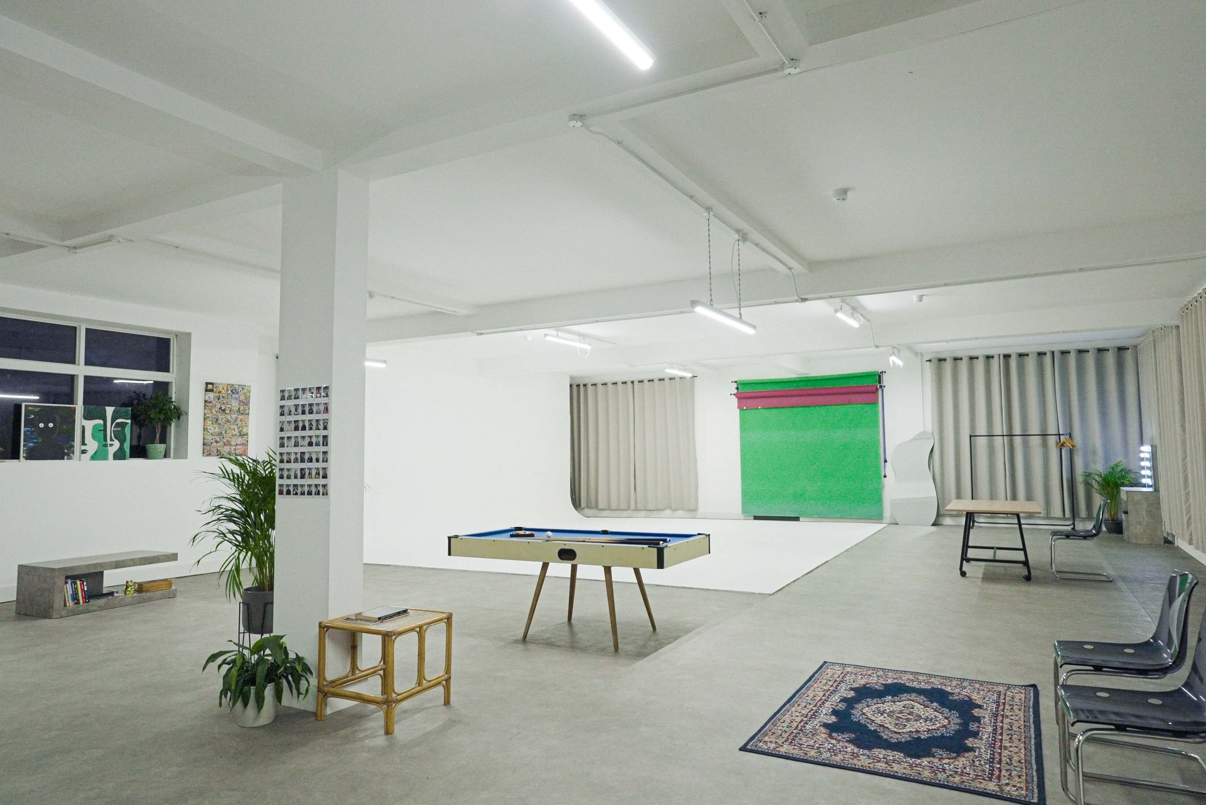 The image shows a spacious studio with a pool table, a small seating area, and a green backdrop for photography, illuminated by bright overhead lights.