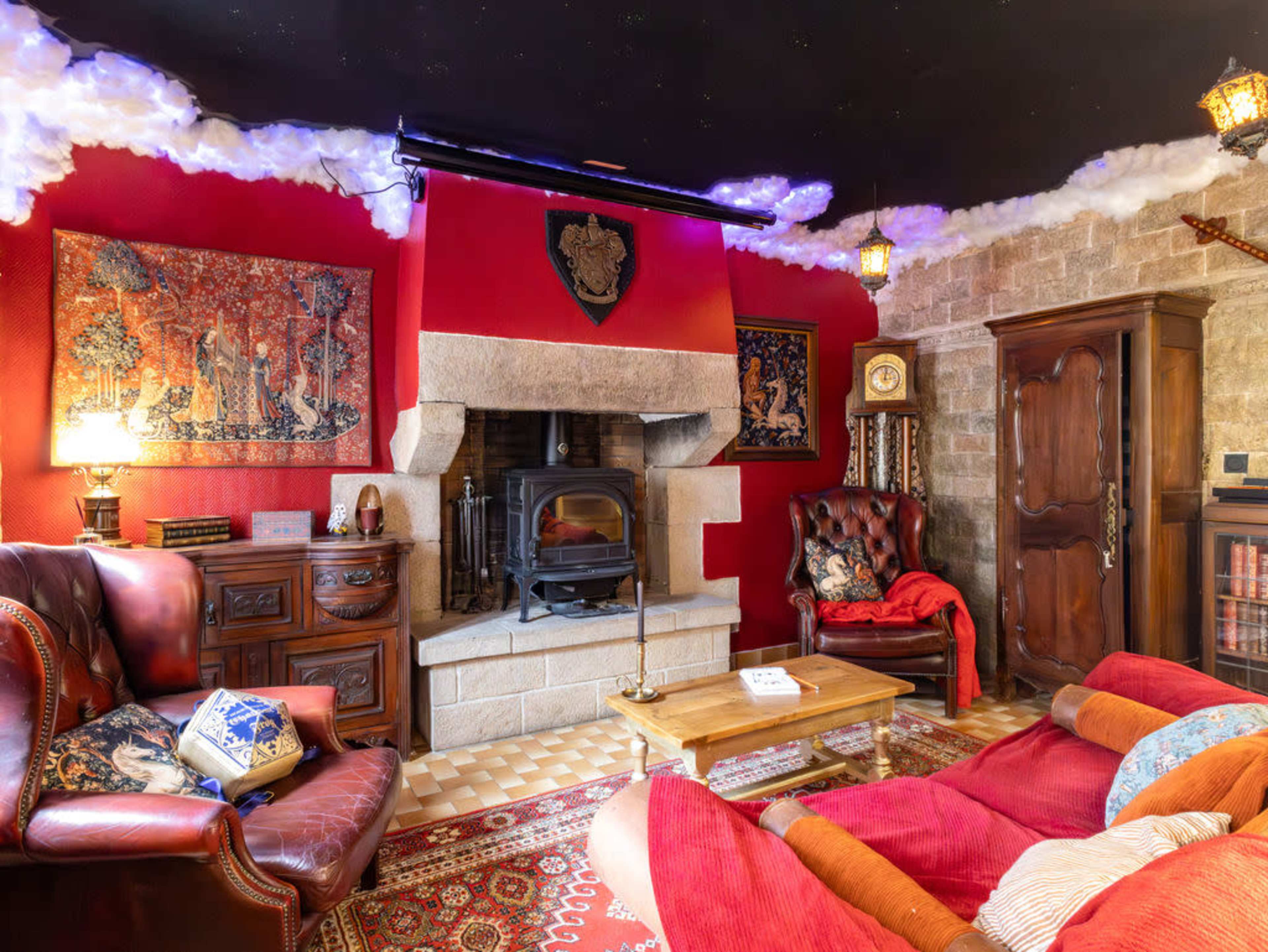 The image shows a cozy living room with a stone fireplace, red walls, ornate decorations, and comfortable seating including a leather armchair and a plush sofa.