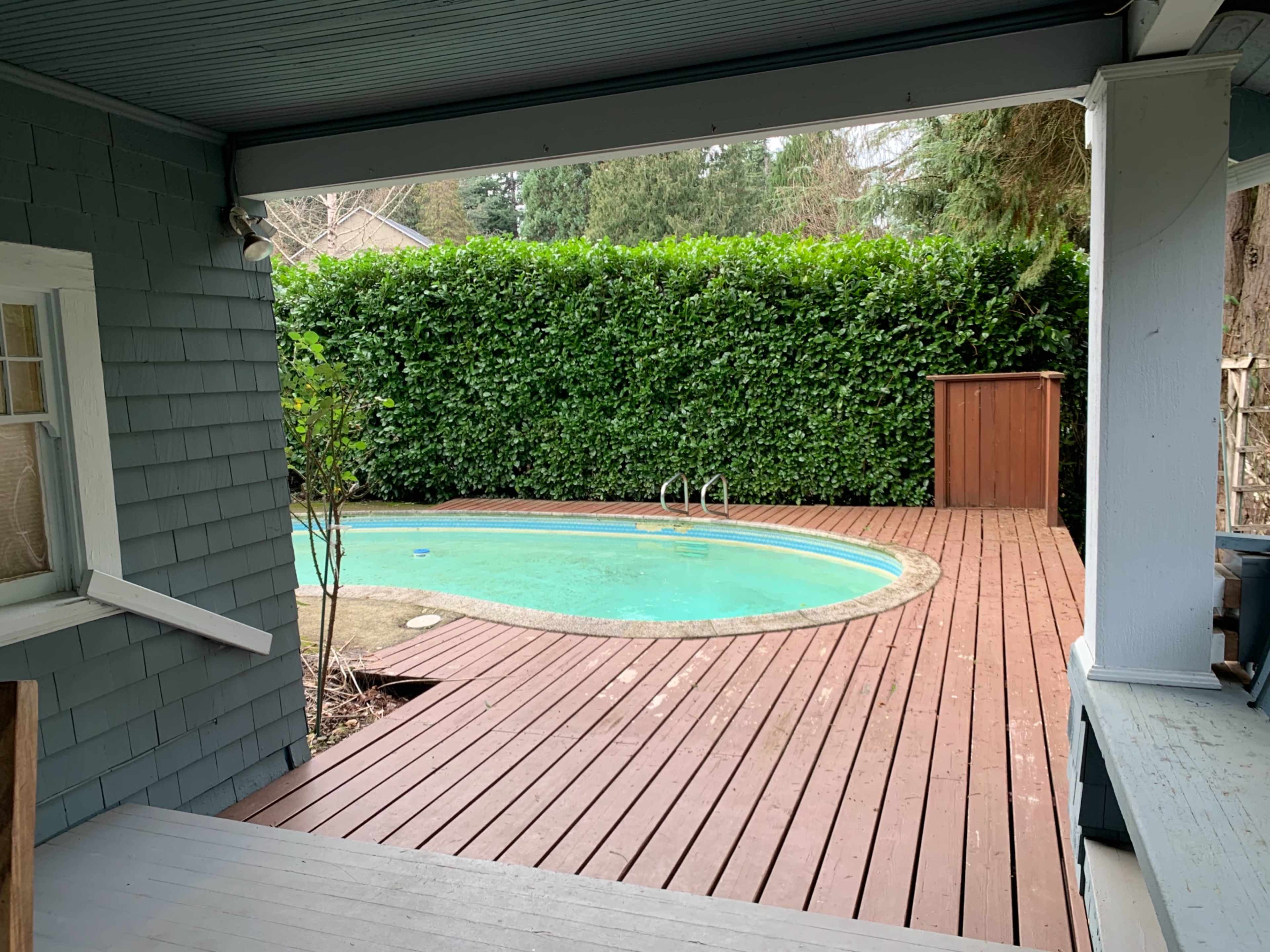 A view from a covered porch overlooking a circular pool surrounded by a wooden deck and a hedge.