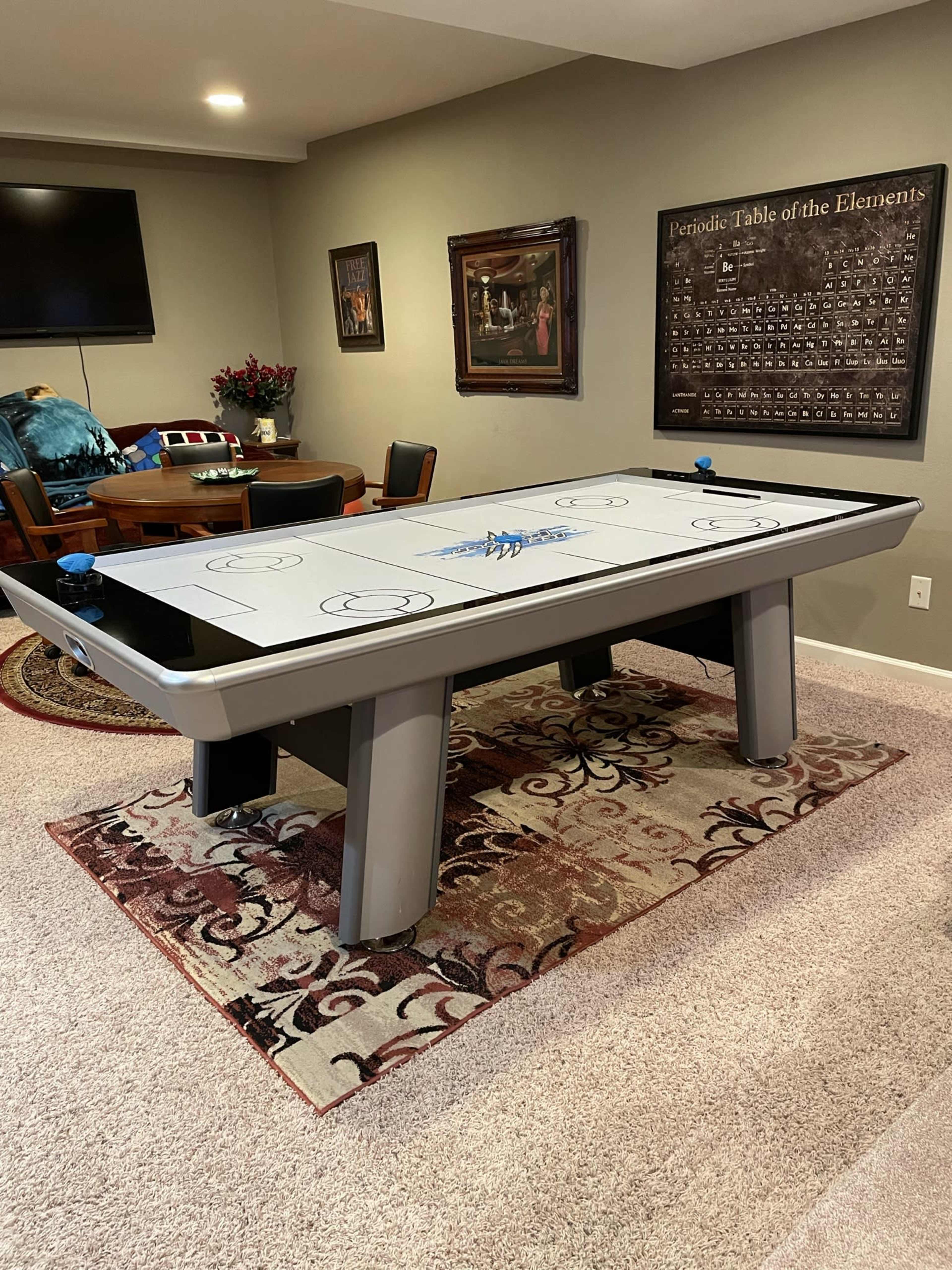 A large air hockey table set on a patterned rug in a room with a flat-screen television and a dining table in the background.