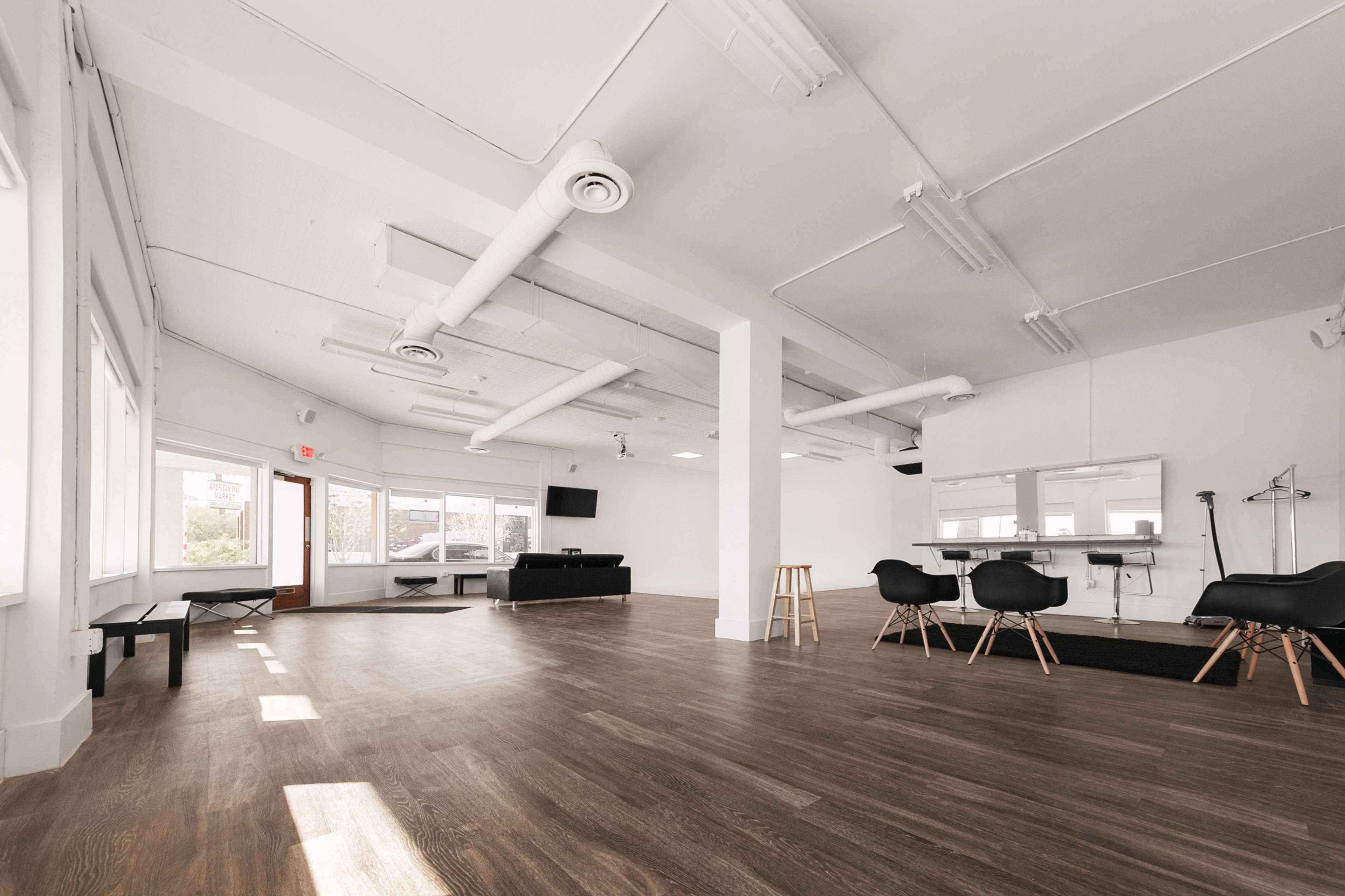 A spacious, bright room with wooden flooring, large windows, and minimalistic furnishings, including a long table and several black chairs.