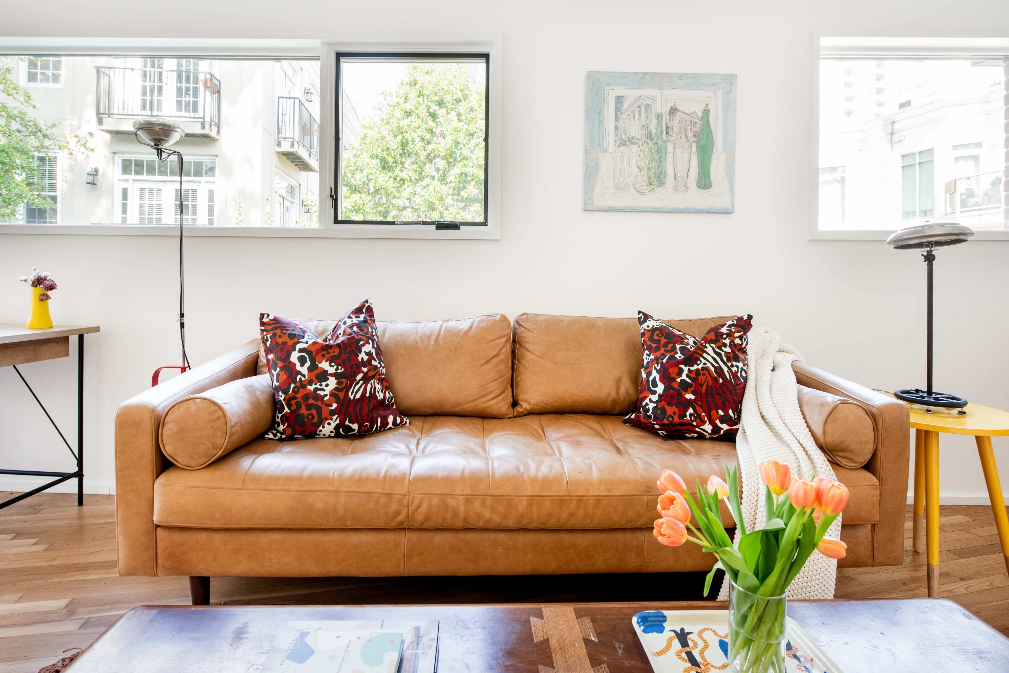 A light-filled living room features a brown leather sofa with patterned cushions, a wooden table, and a vase of tulips.