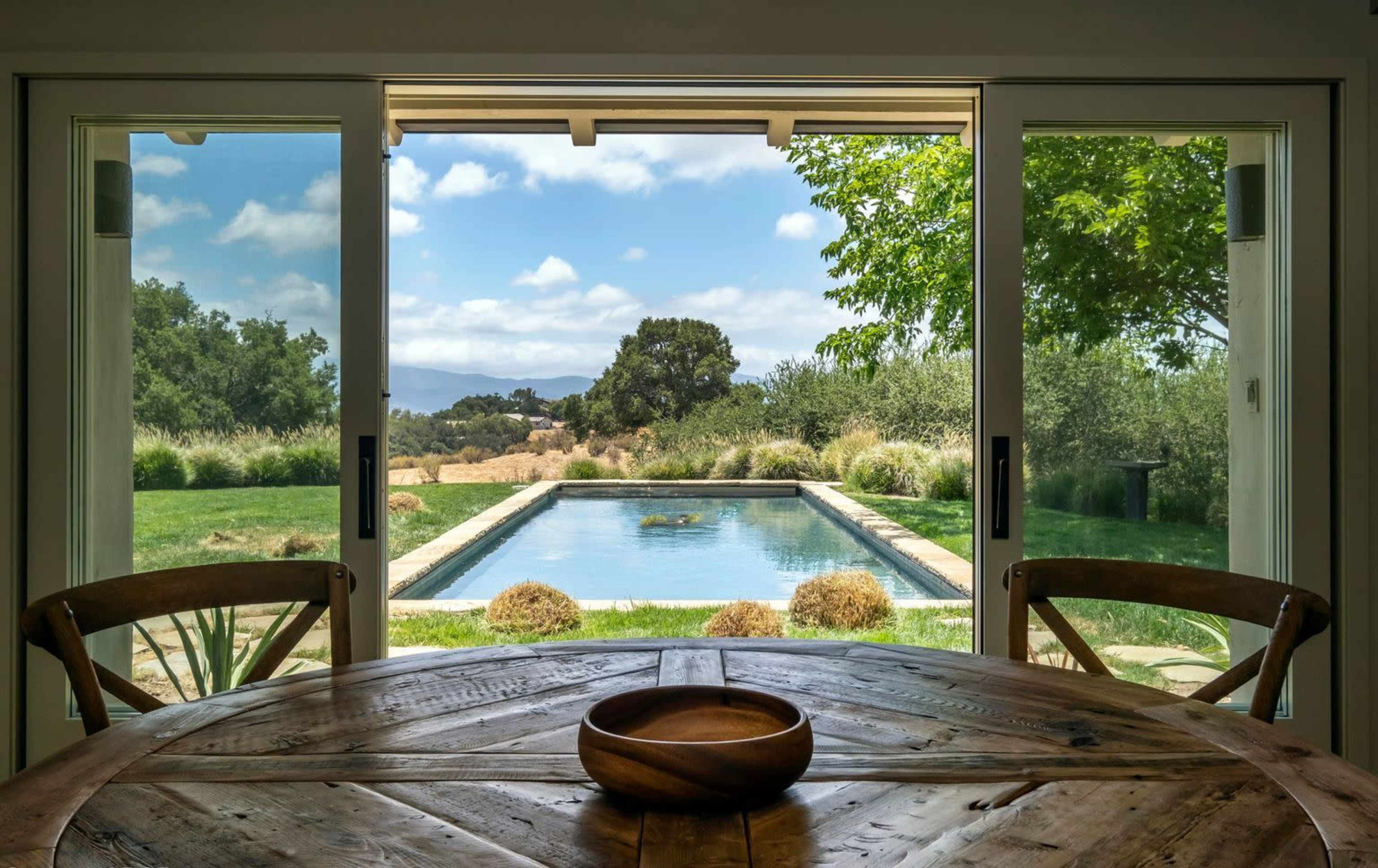A wooden dining table sits in front of large open doors, revealing a pool and landscaped backyard view with trees and clouds in the sky.
