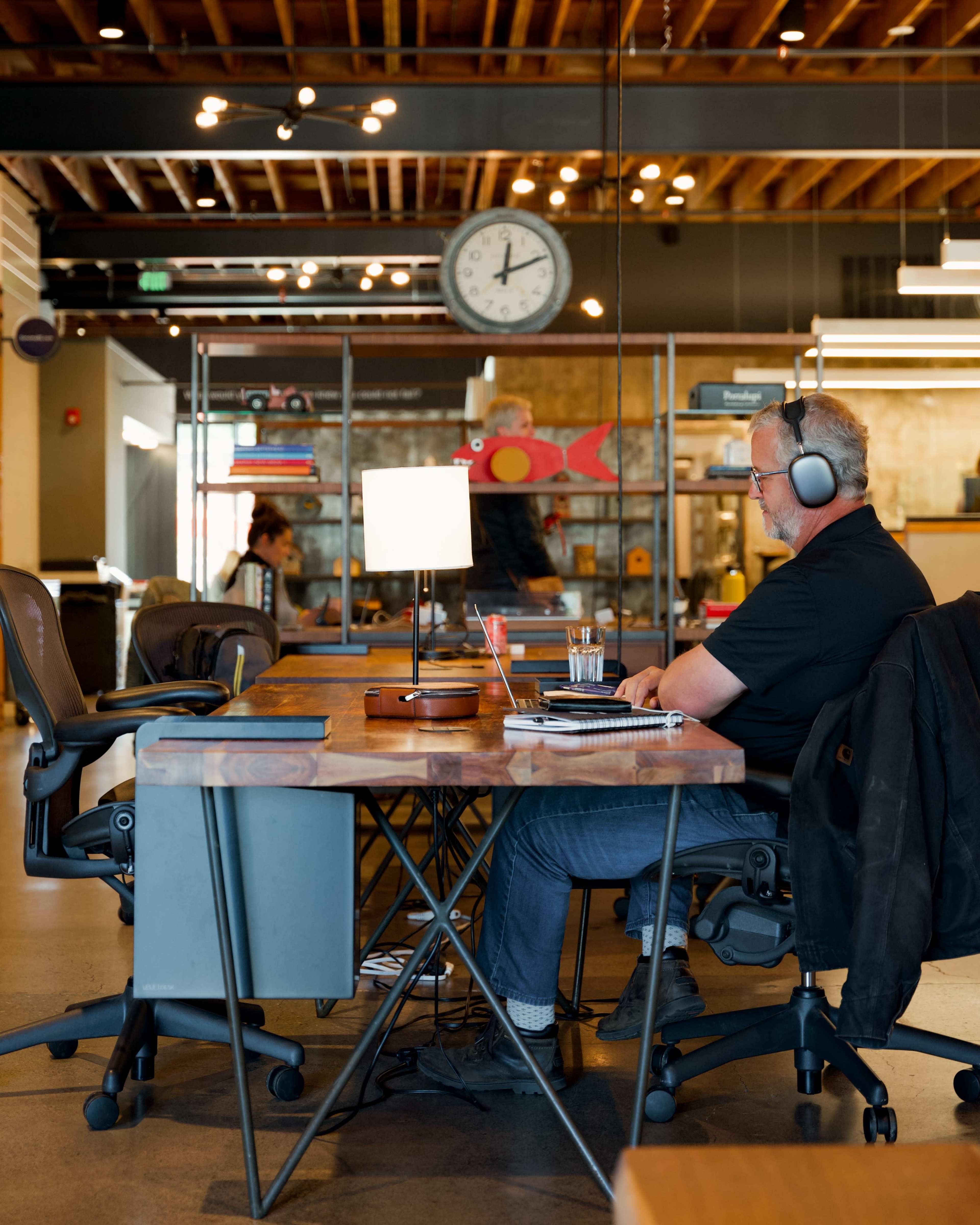 A man wearing headphones sits at a wooden desk in a modern workspace, while a clock hangs on the wall and other people are visible in the background.