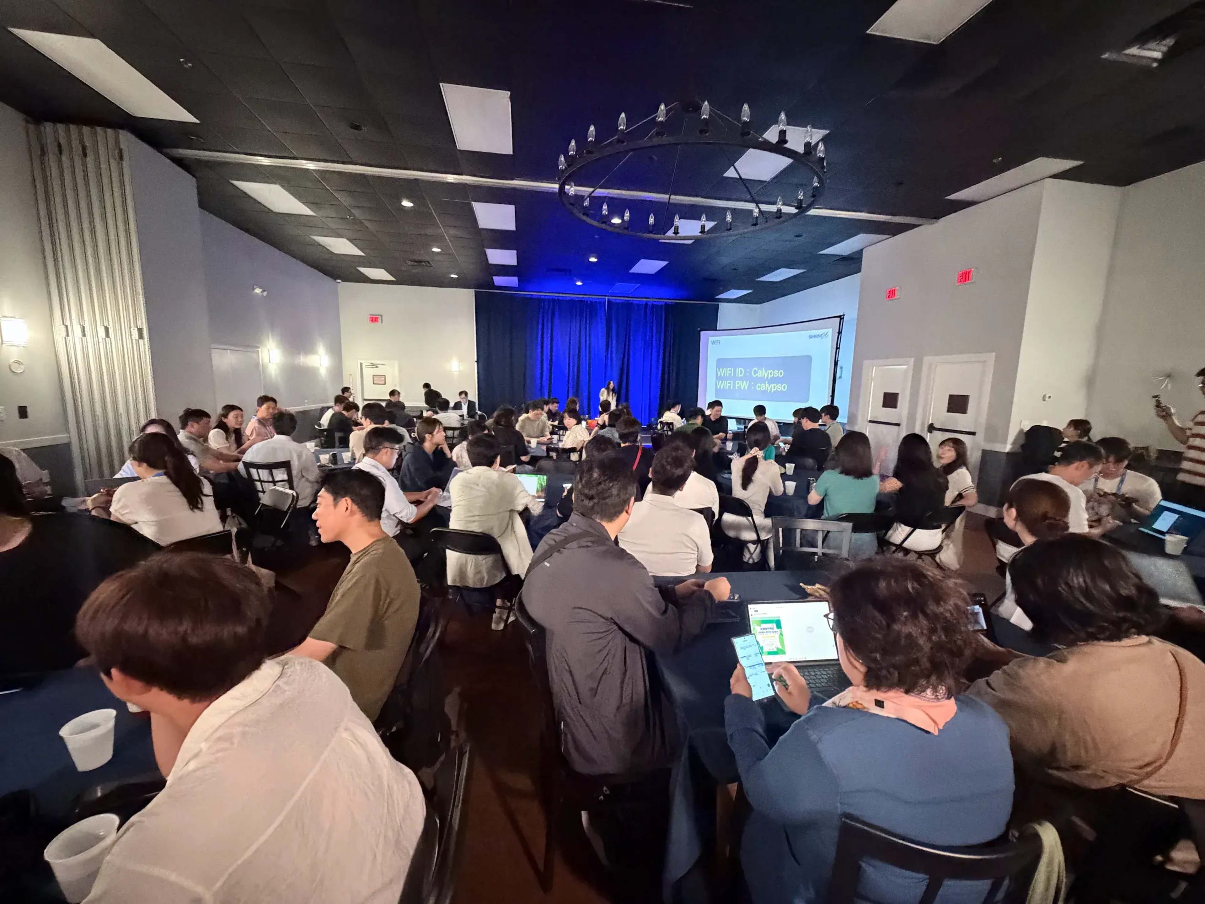 A large group of people is seated at tables in a dimly lit room, with a speaker presenting in front of a screen displaying information.