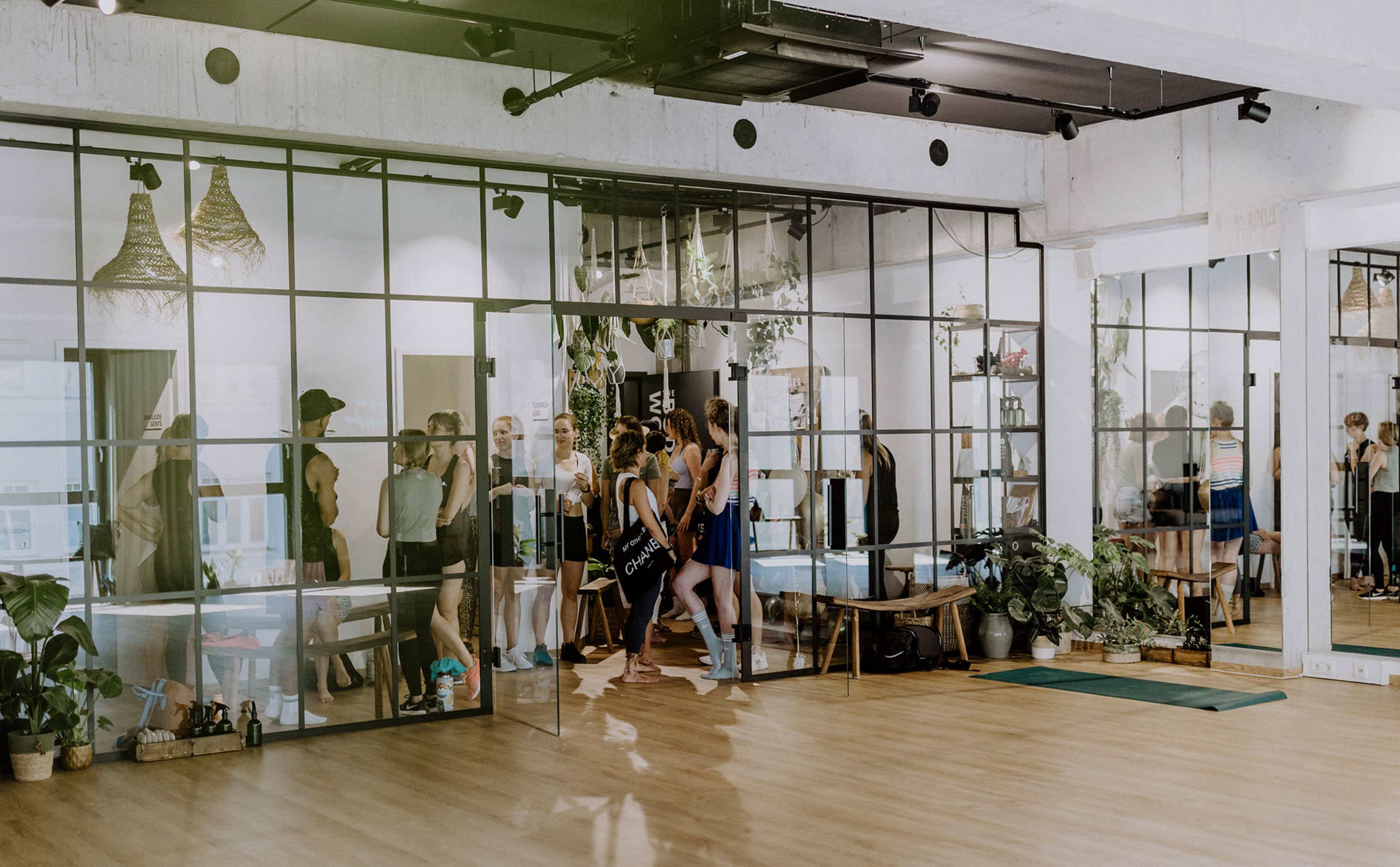 A group of people is engaged in a fitness class inside a modern studio with large glass walls and wooden flooring, surrounded by indoor plants.