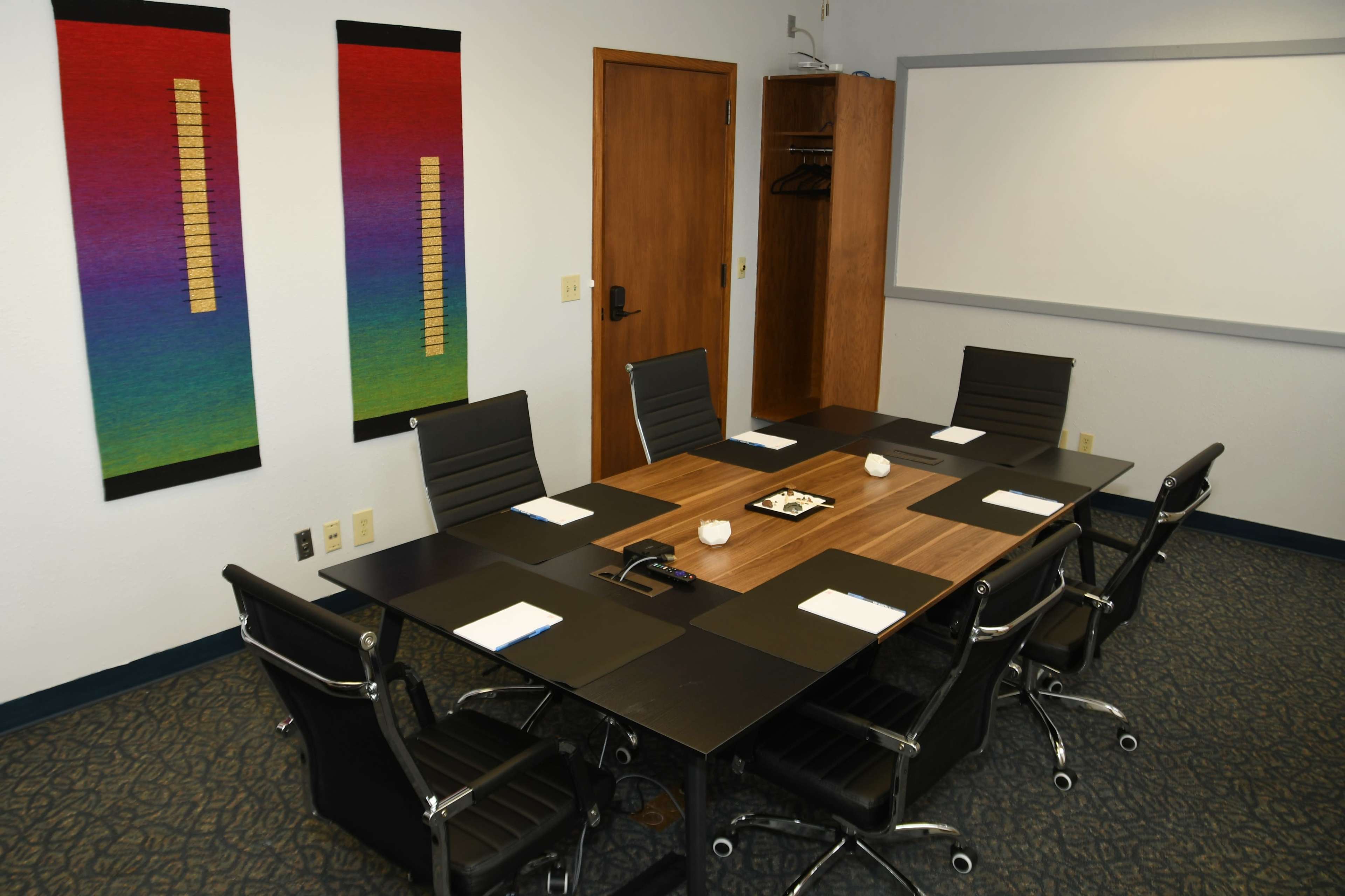 A modern conference room features a large black table surrounded by ergonomic chairs, with colorful wall art and a whiteboard in the background.