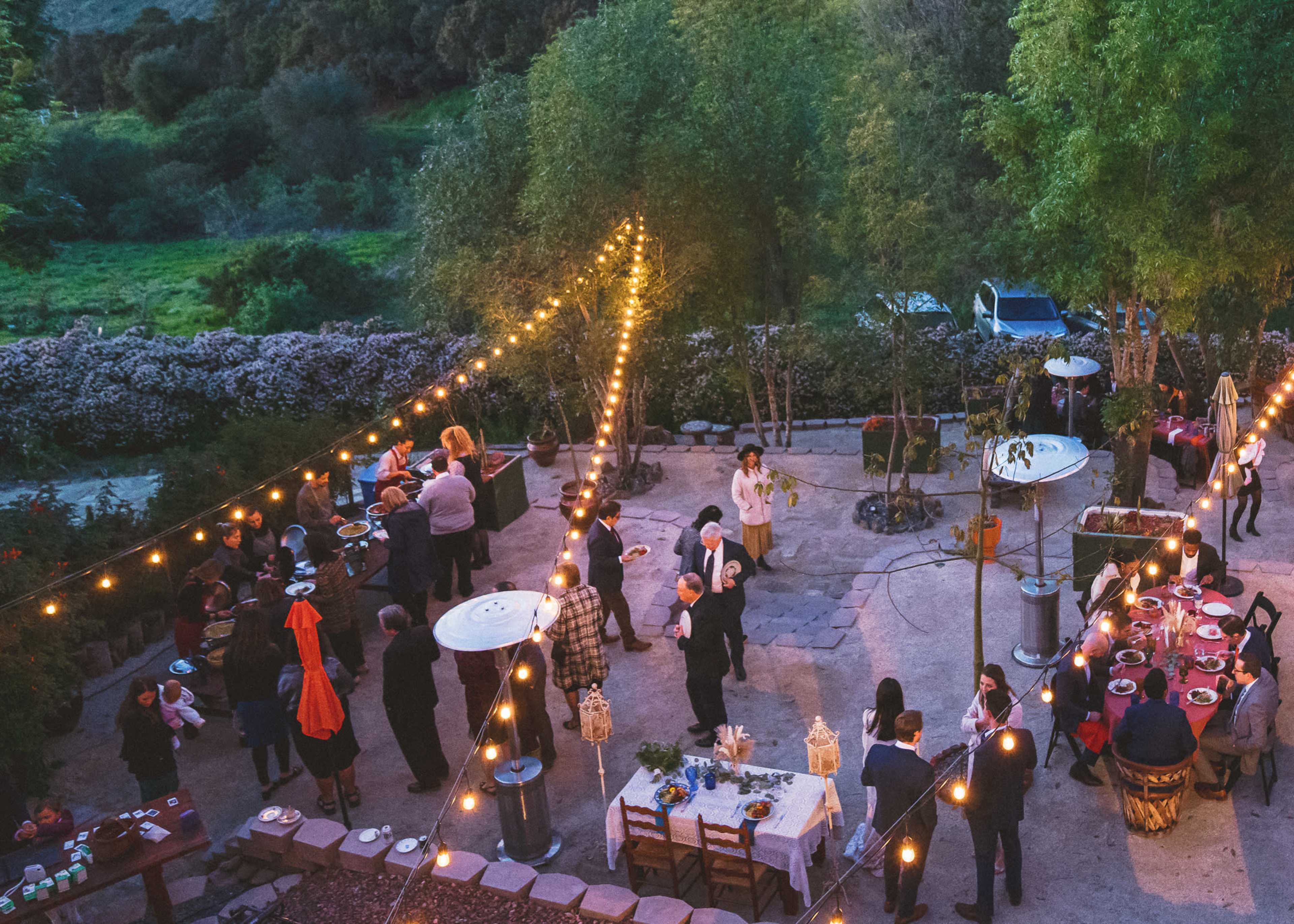 A gathering takes place outdoors in a landscaped area, illuminated by string lights, with guests mingling around tables set for a meal.