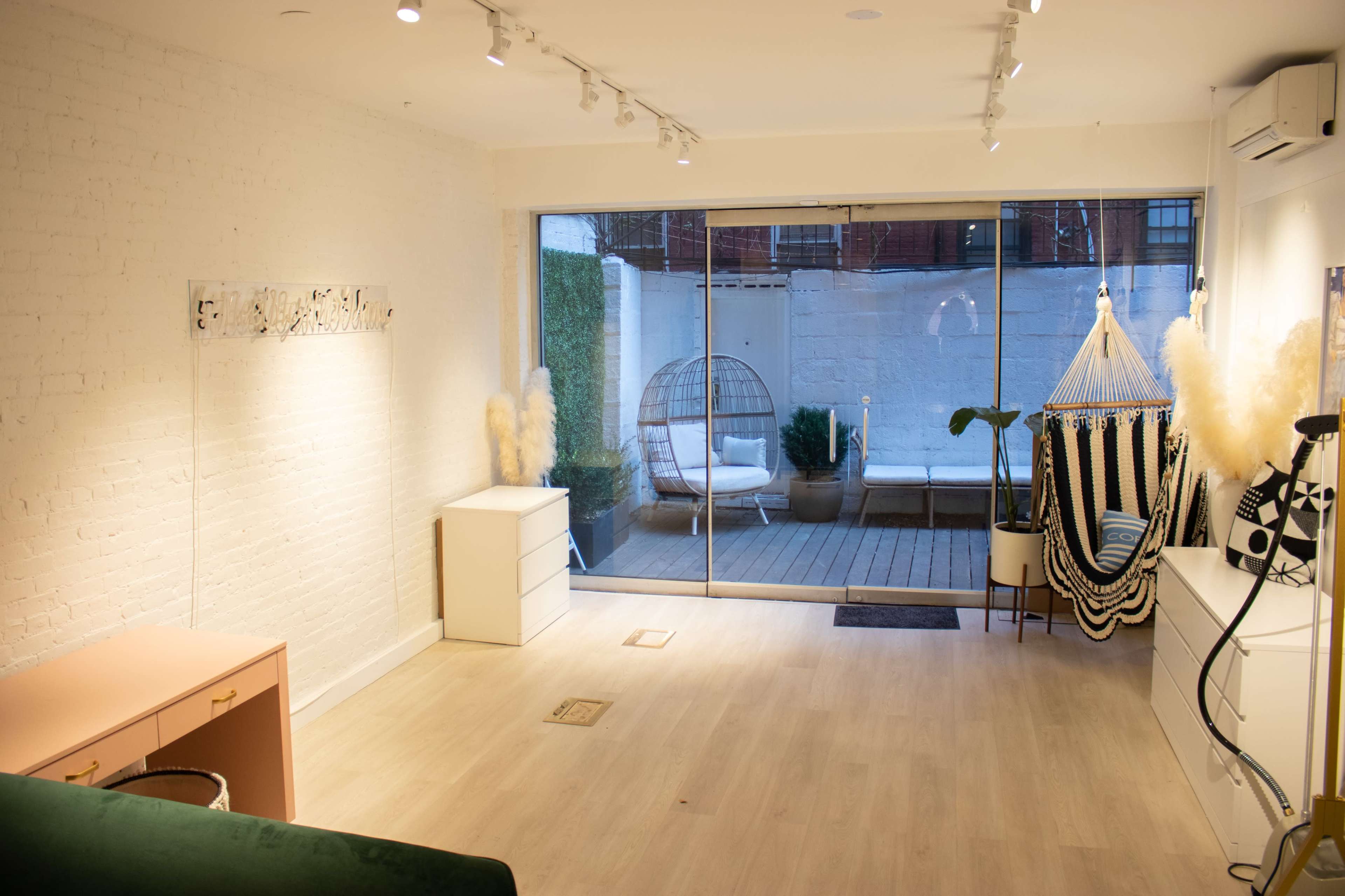 A minimalist interior space with light-colored walls, wooden flooring, and a large sliding glass door leading to a patio containing furniture and plants.
