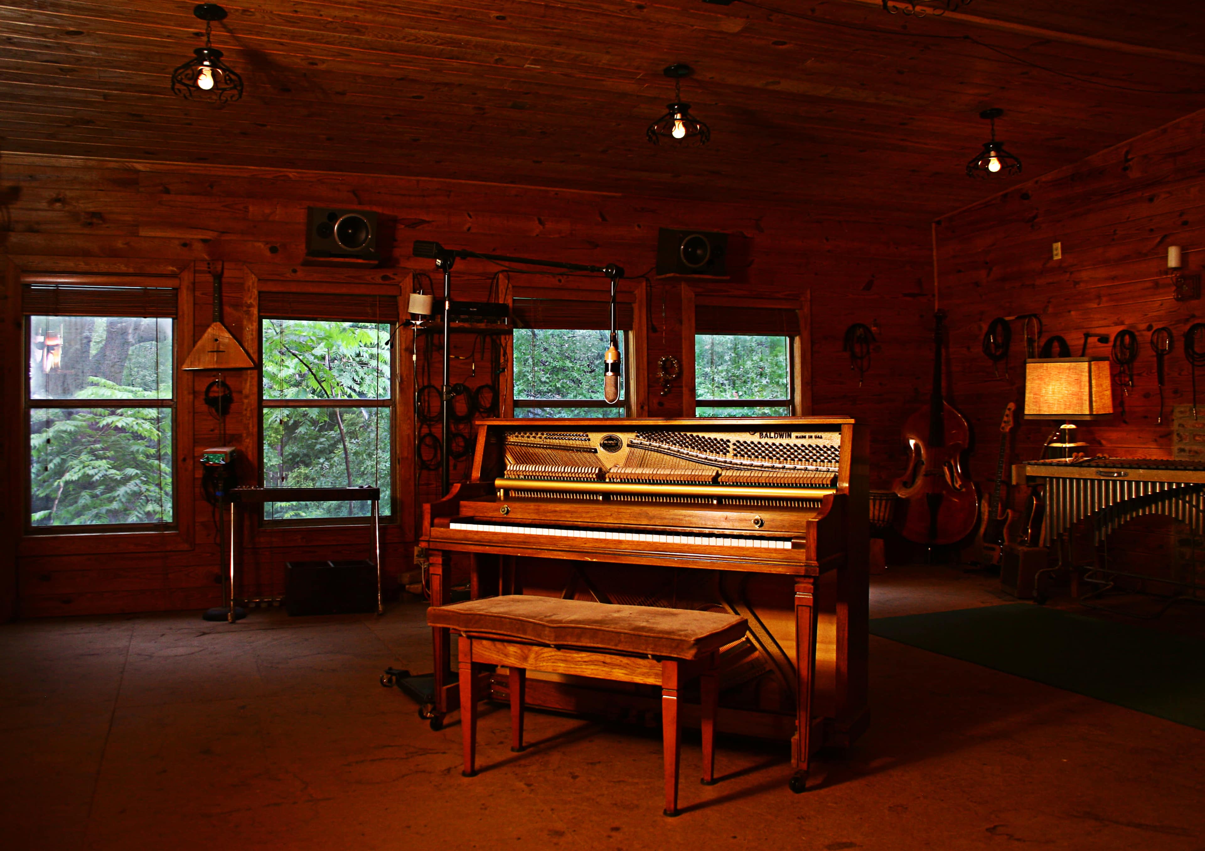 A wooden piano with a matching bench sits in a cozy, rustic room with wooden walls and multiple windows.