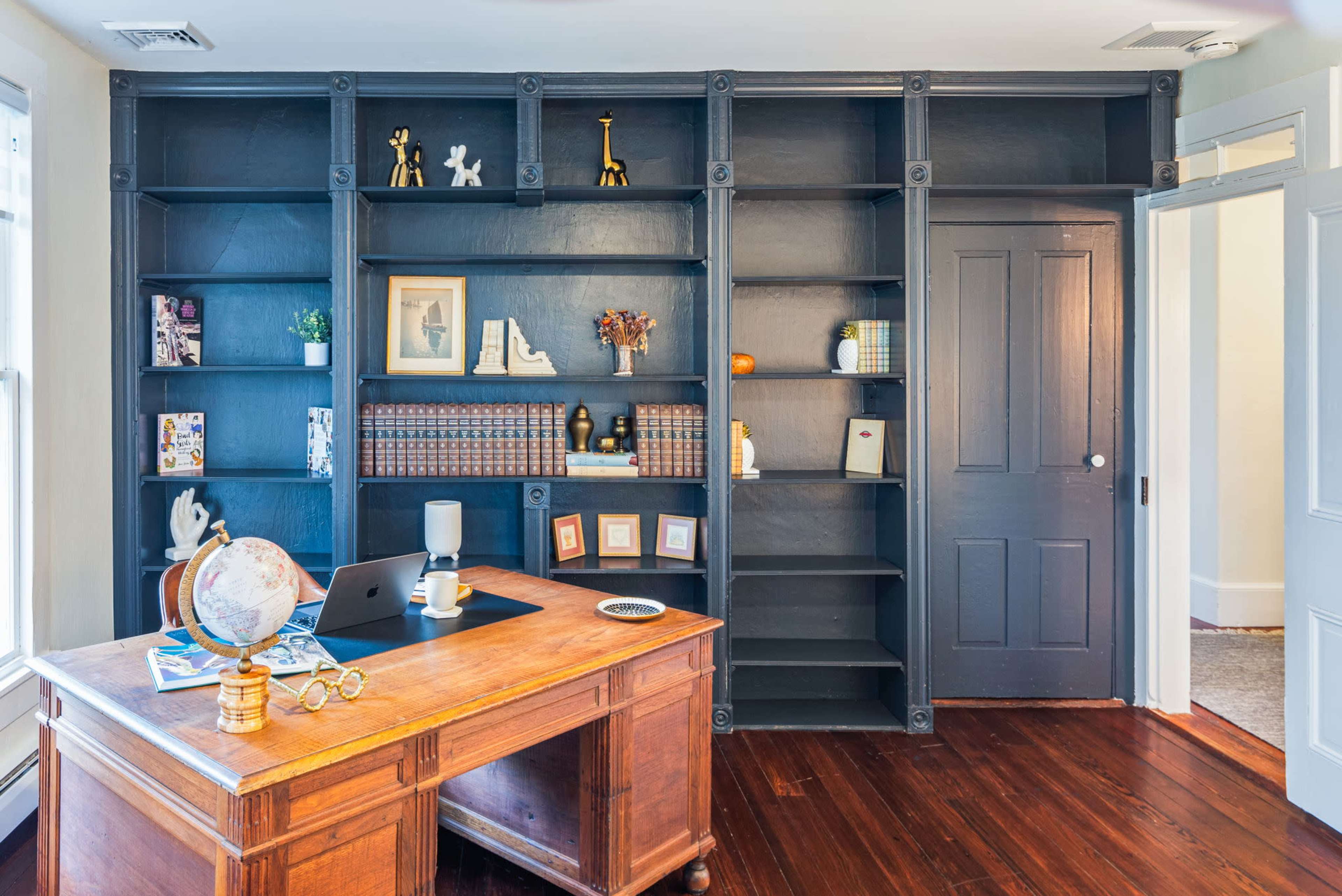 The image shows a home office featuring a wooden desk with a globe, a computer, and bookshelf units filled with decorative items and books against a dark wall.