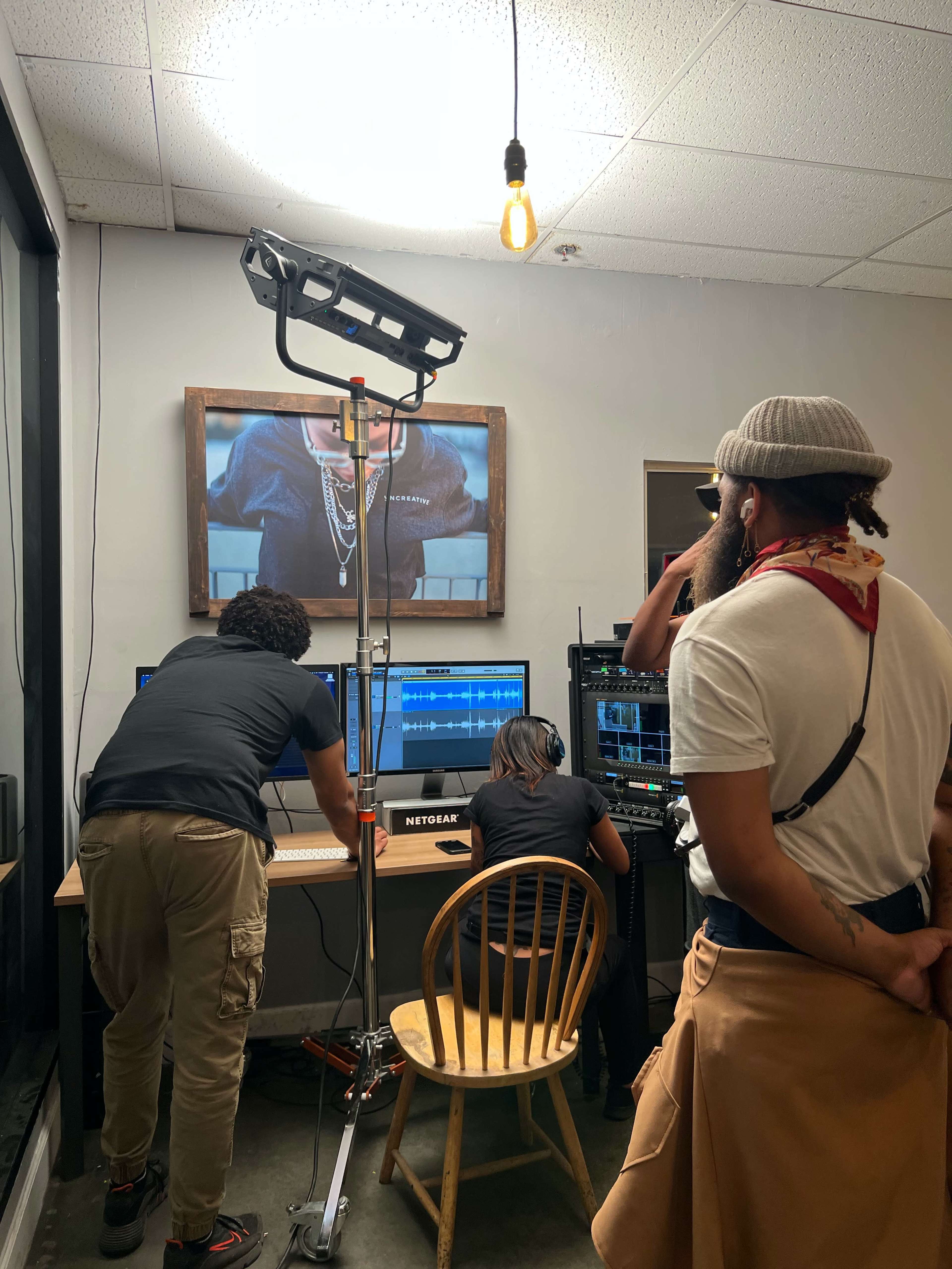 Three individuals work at computer stations with editing software while a fourth person observes, in a dimly lit room with a large screen showing video footage in the background.
