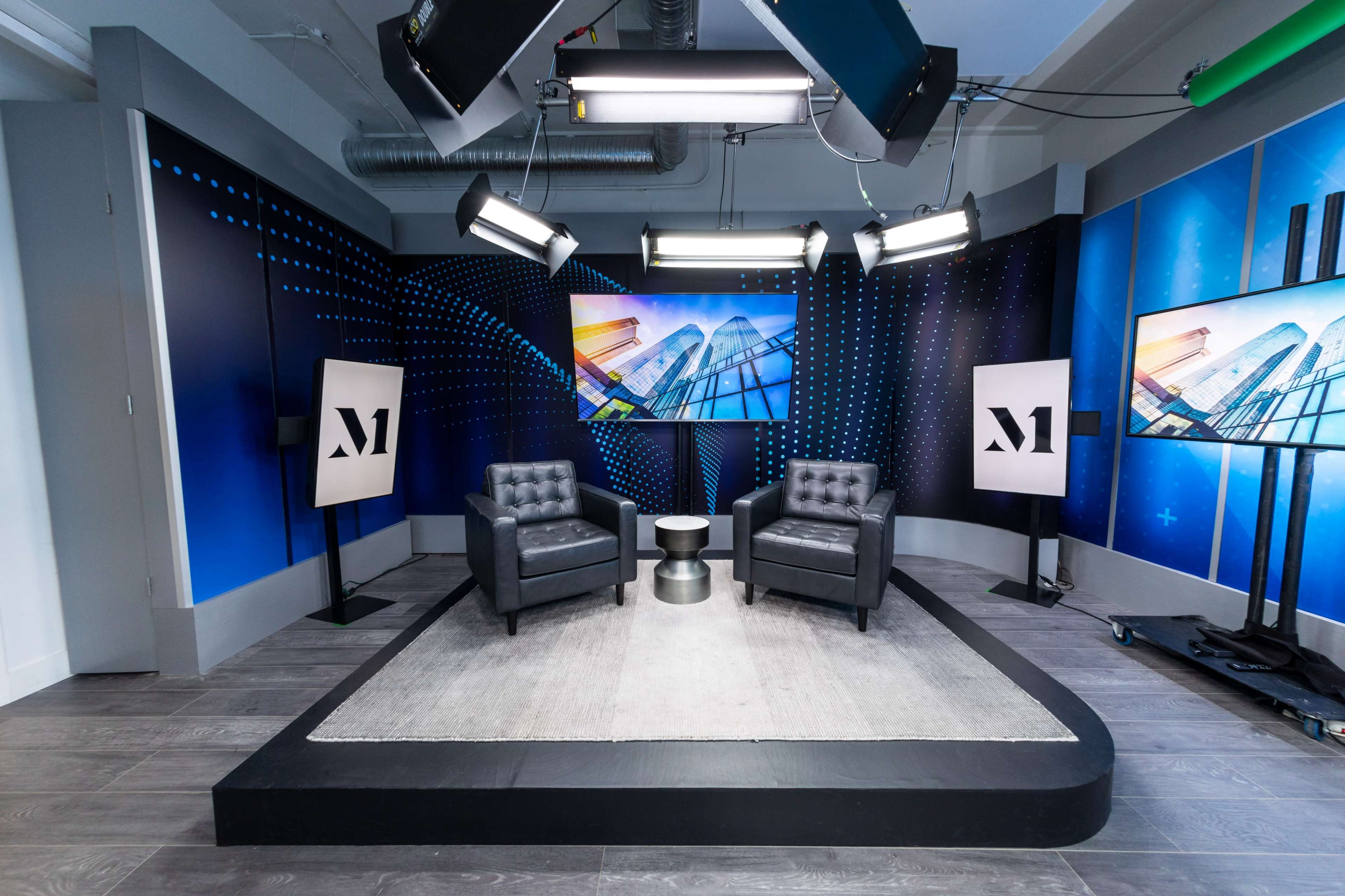A modern studio setup featuring two black chairs facing a circular table, with large screens displaying graphics and studio lighting overhead.