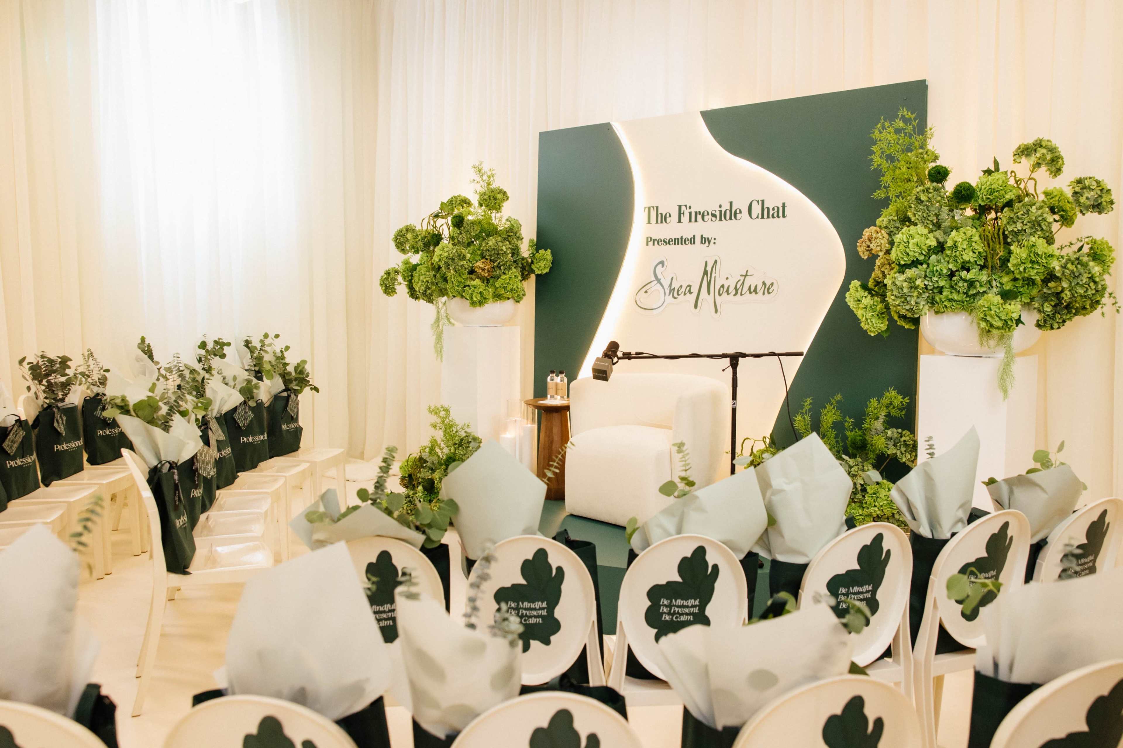 The image shows a neatly arranged event space for a fireside chat, featuring white chairs facing a backdrop decorated with greenery and a branded logo.