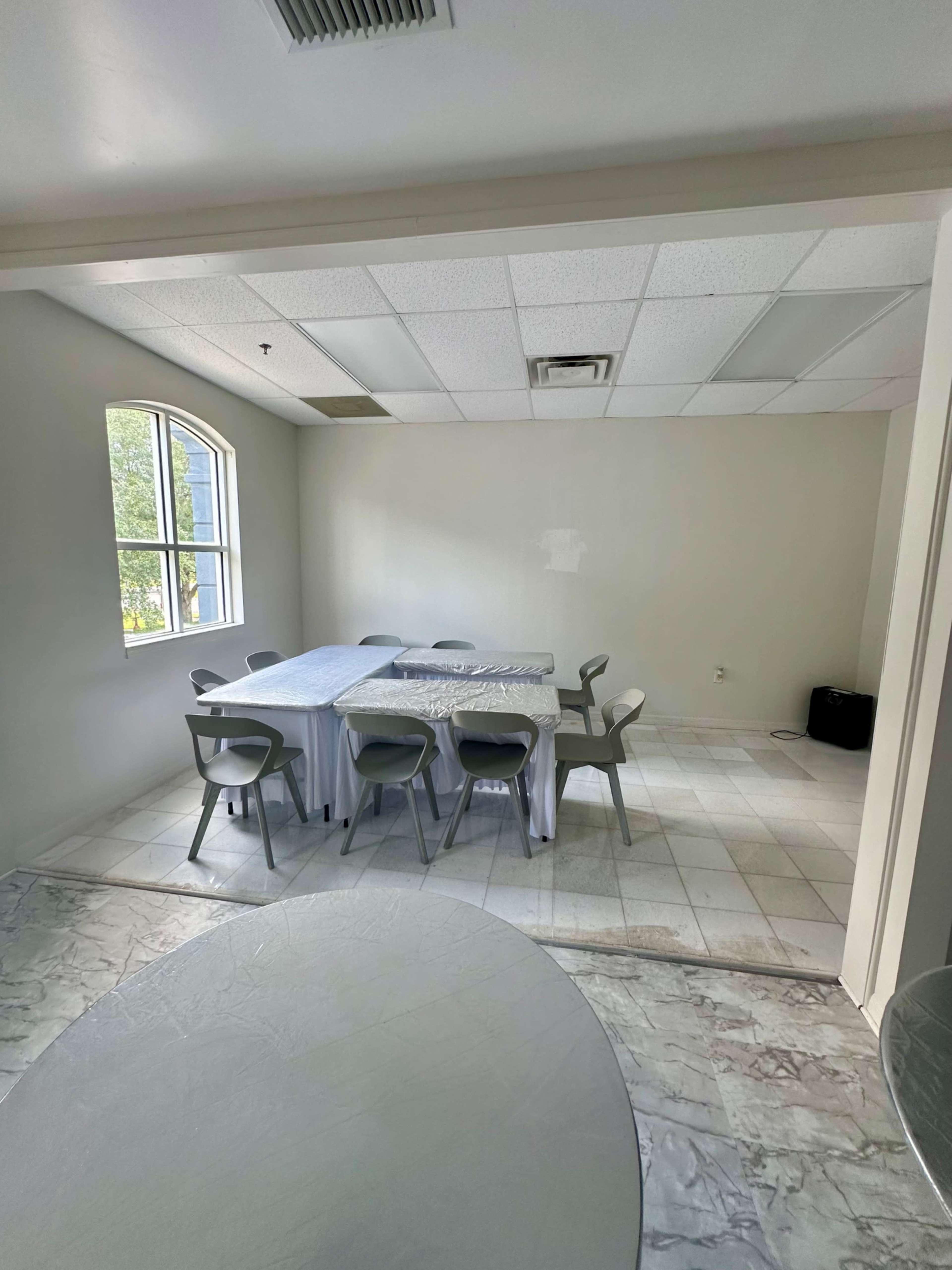 A simple, sparsely furnished room with a rectangular table covered by a white tablecloth and surrounded by eight chairs.