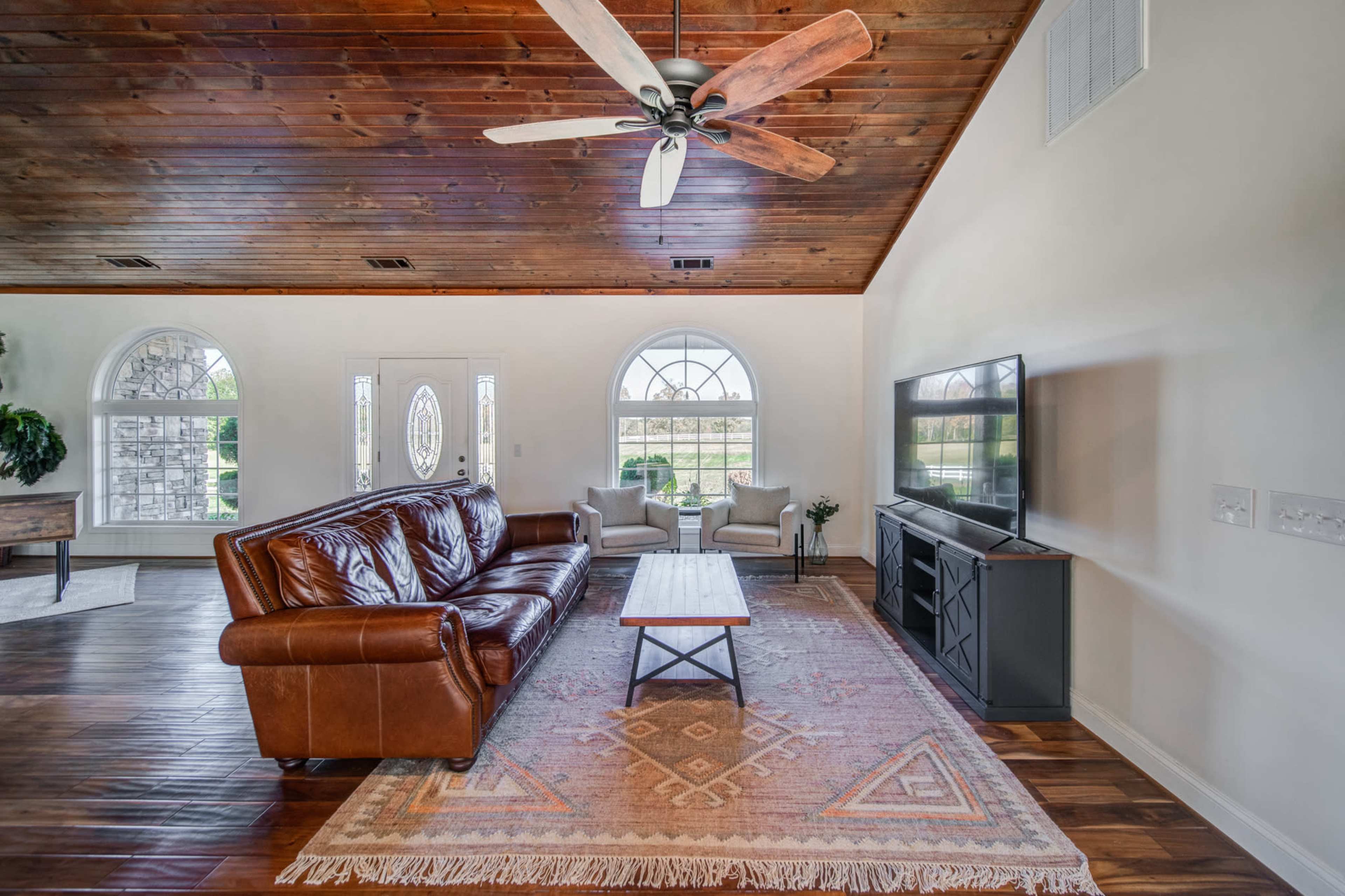 The living room features a brown leather sofa, a wooden coffee table, and a large television against a wall, with two armchairs and arched windows providing natural light.