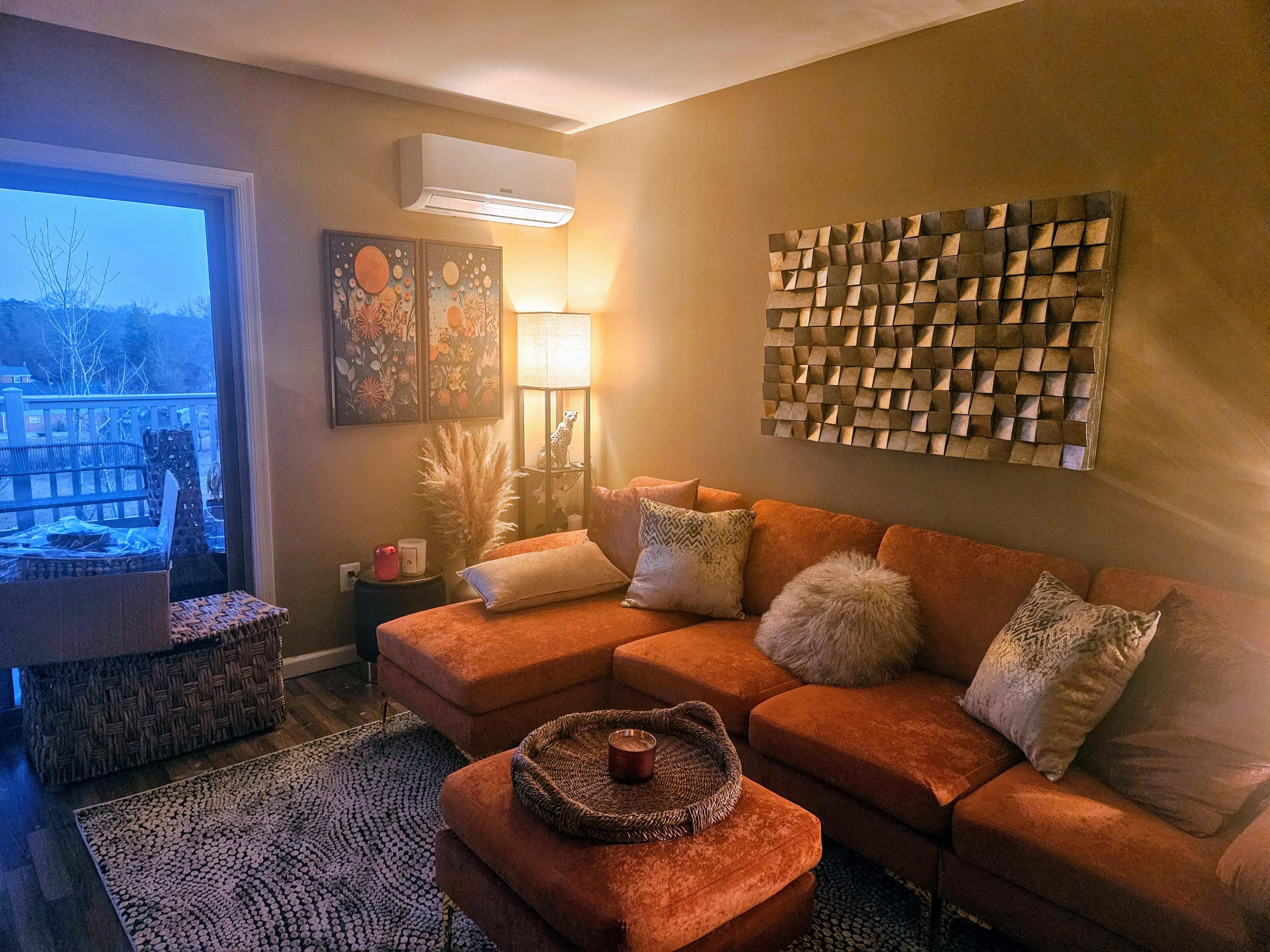 The image shows a cozy living room featuring an orange sectional sofa, a decorative wall hanging, and a window overlooking a dimly lit outdoor area.