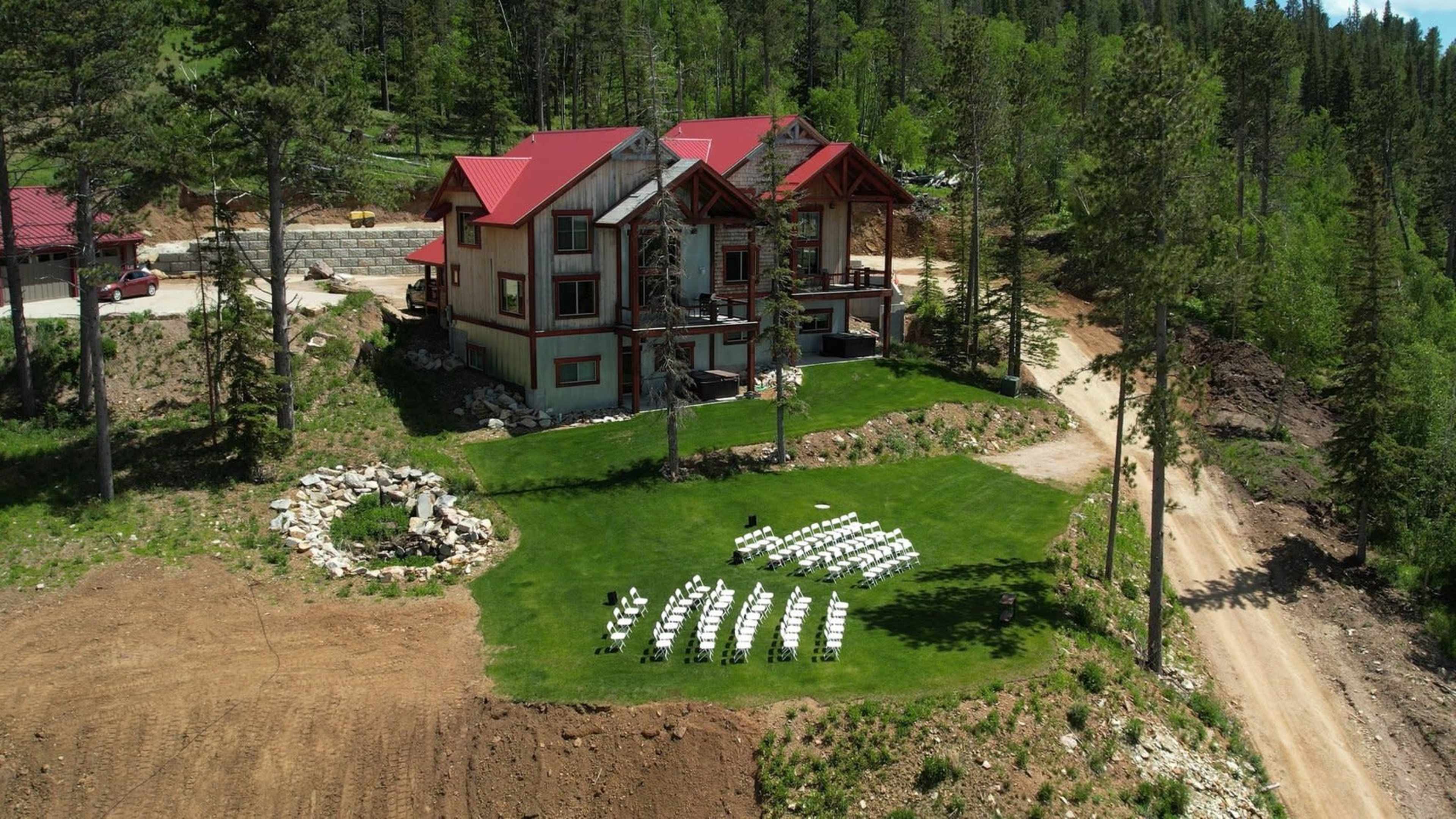 A large house with a red roof is surrounded by trees, and a circular seating arrangement of white chairs sits on a grassy area nearby.