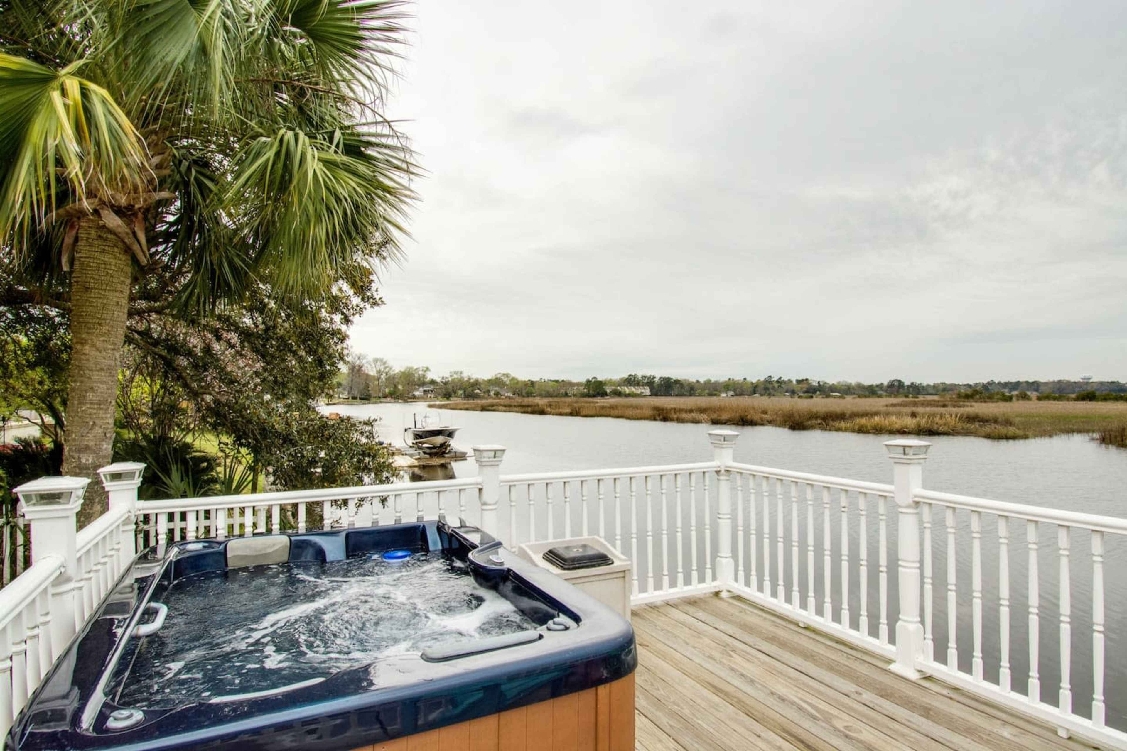 A hot tub sits on a deck overlooking a river with grassy banks and a boat in the distance, framed by palm trees.