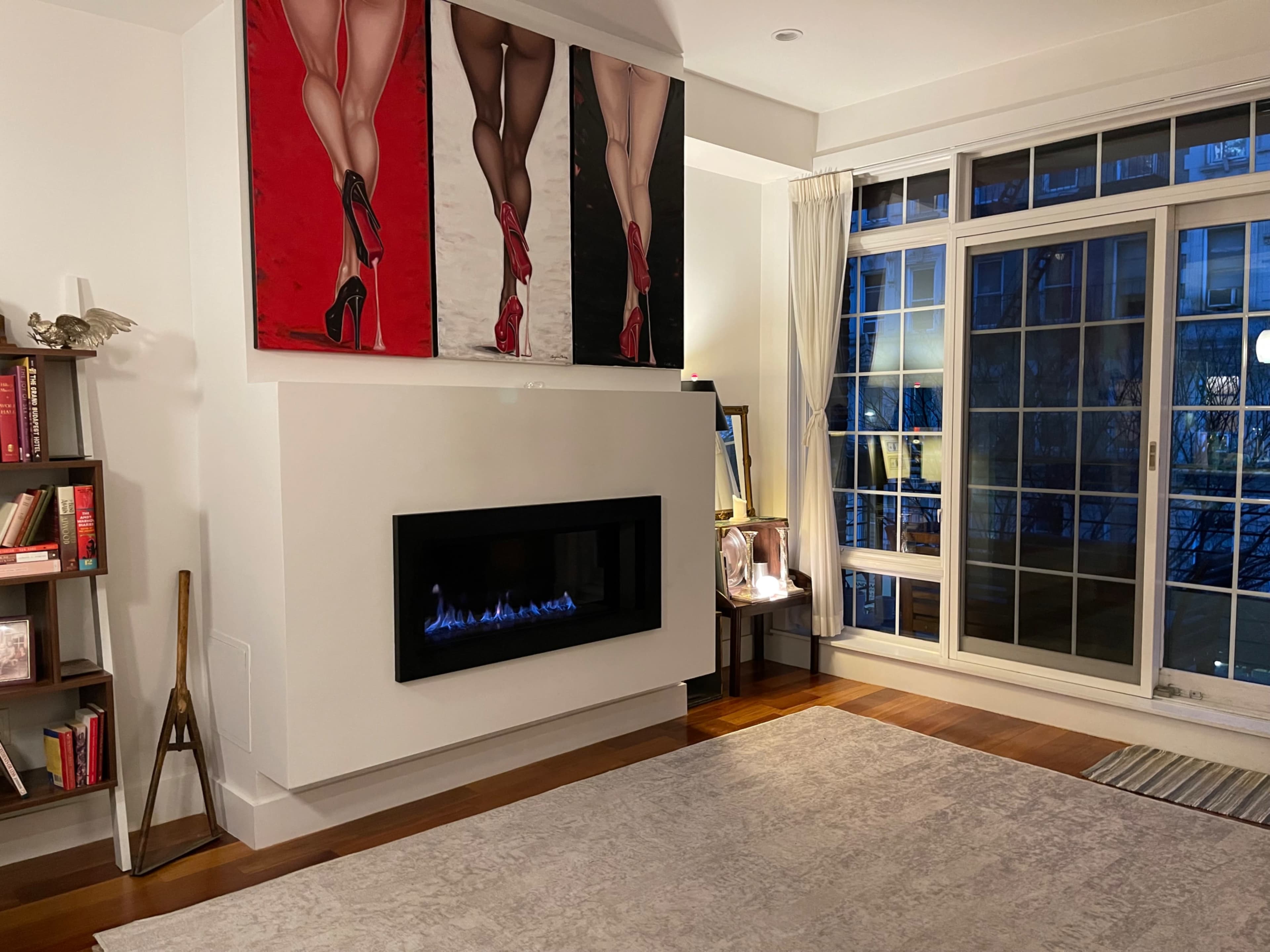 A modern living room with a wall-mounted fireplace, a large artwork of legs in high heels above it, and windows overlooking an outdoor area.