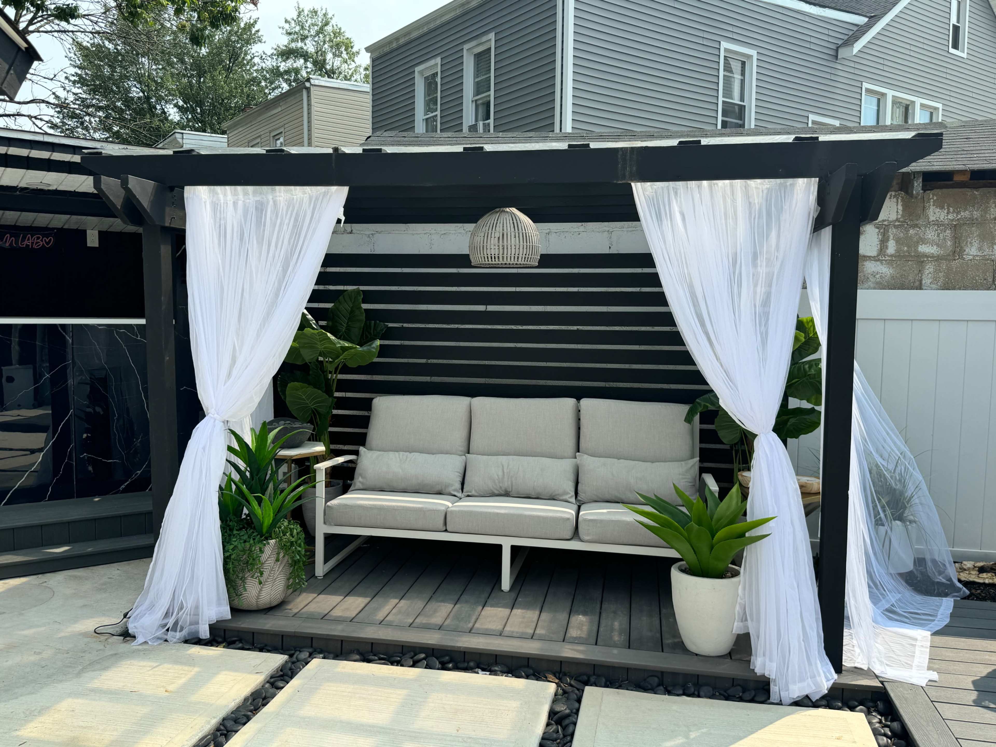 A shaded outdoor seating area features a gray sofa flanked by potted plants and draped white curtains.