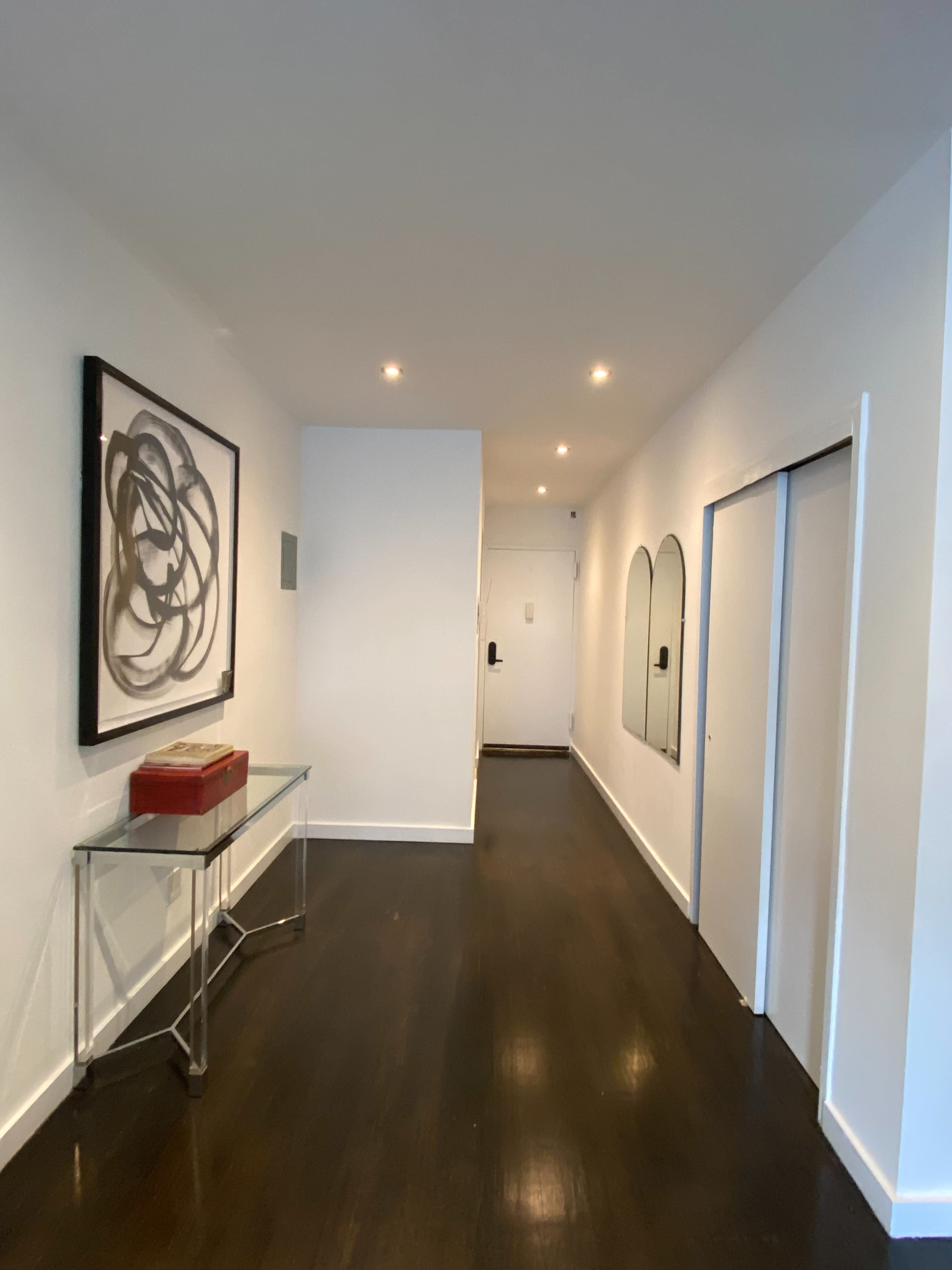 The image shows a narrow hallway with a framed artwork on the left, a glass console table, and mirrored arches above two closet doors, leading to a door at the end.