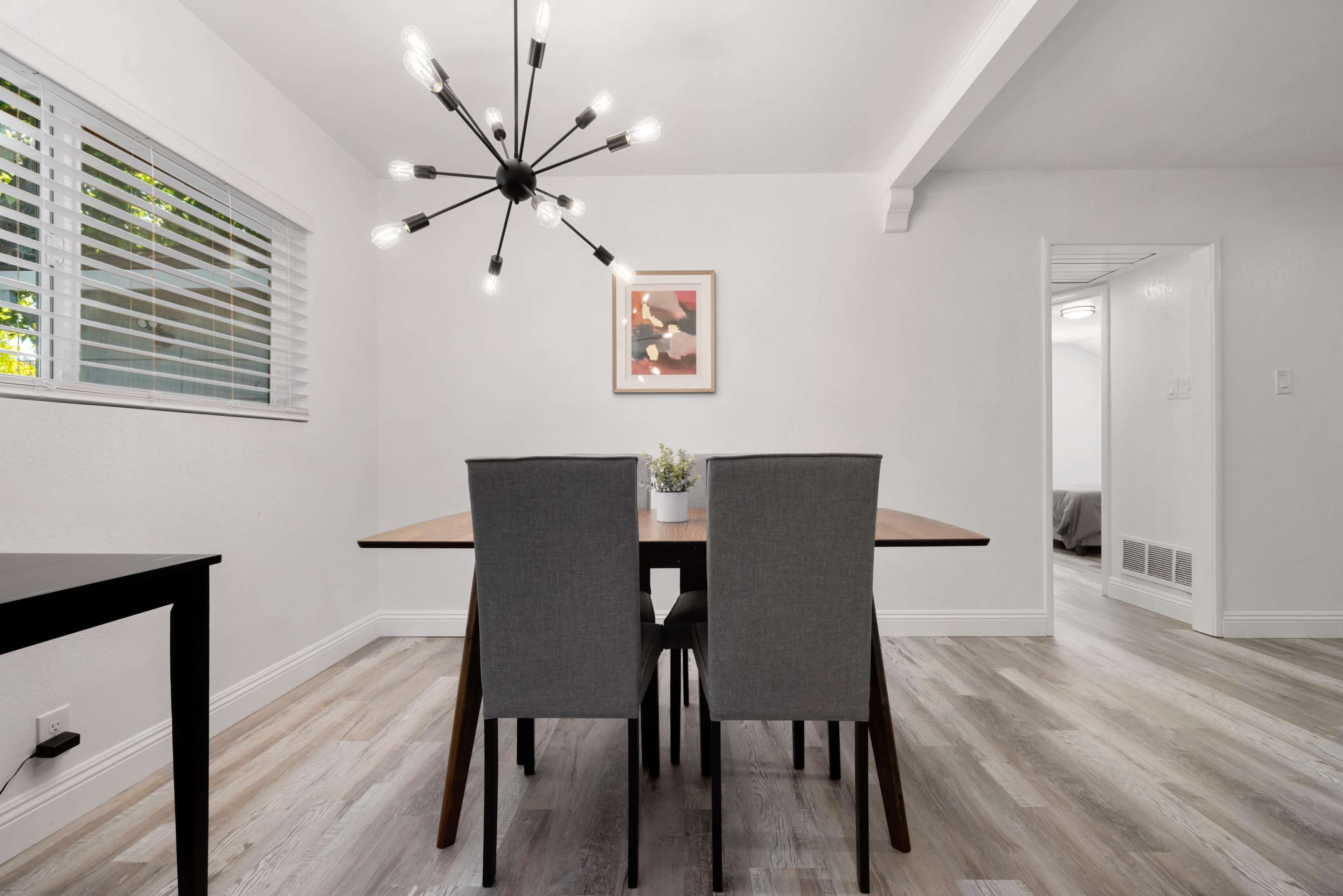 A modern dining area features a wooden table with two gray chairs, overhead lighting, and a window with blinds.