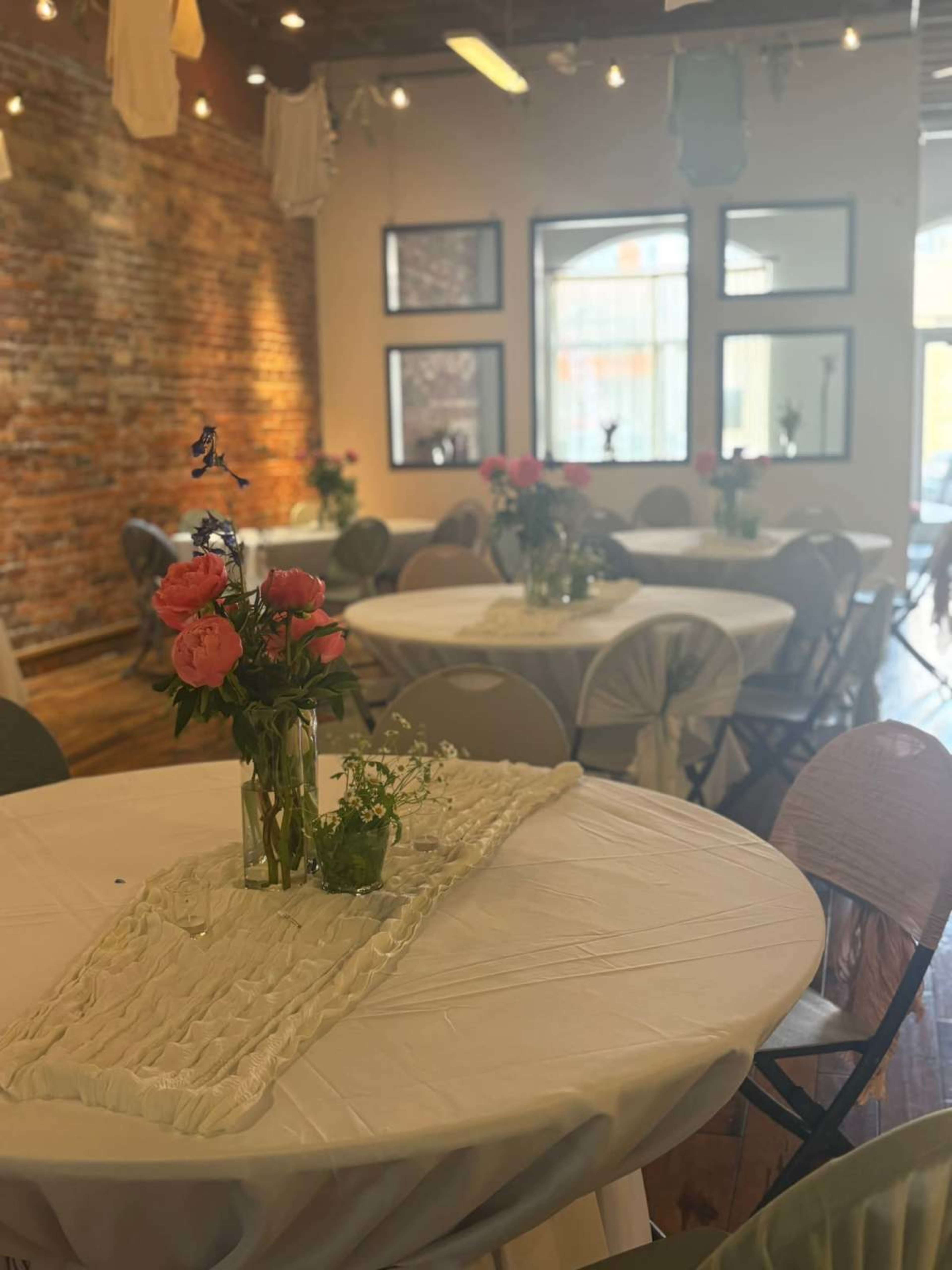 The image shows a dining area set with round tables covered in white tablecloths, decorated with floral centerpieces, and a brick wall in the background.