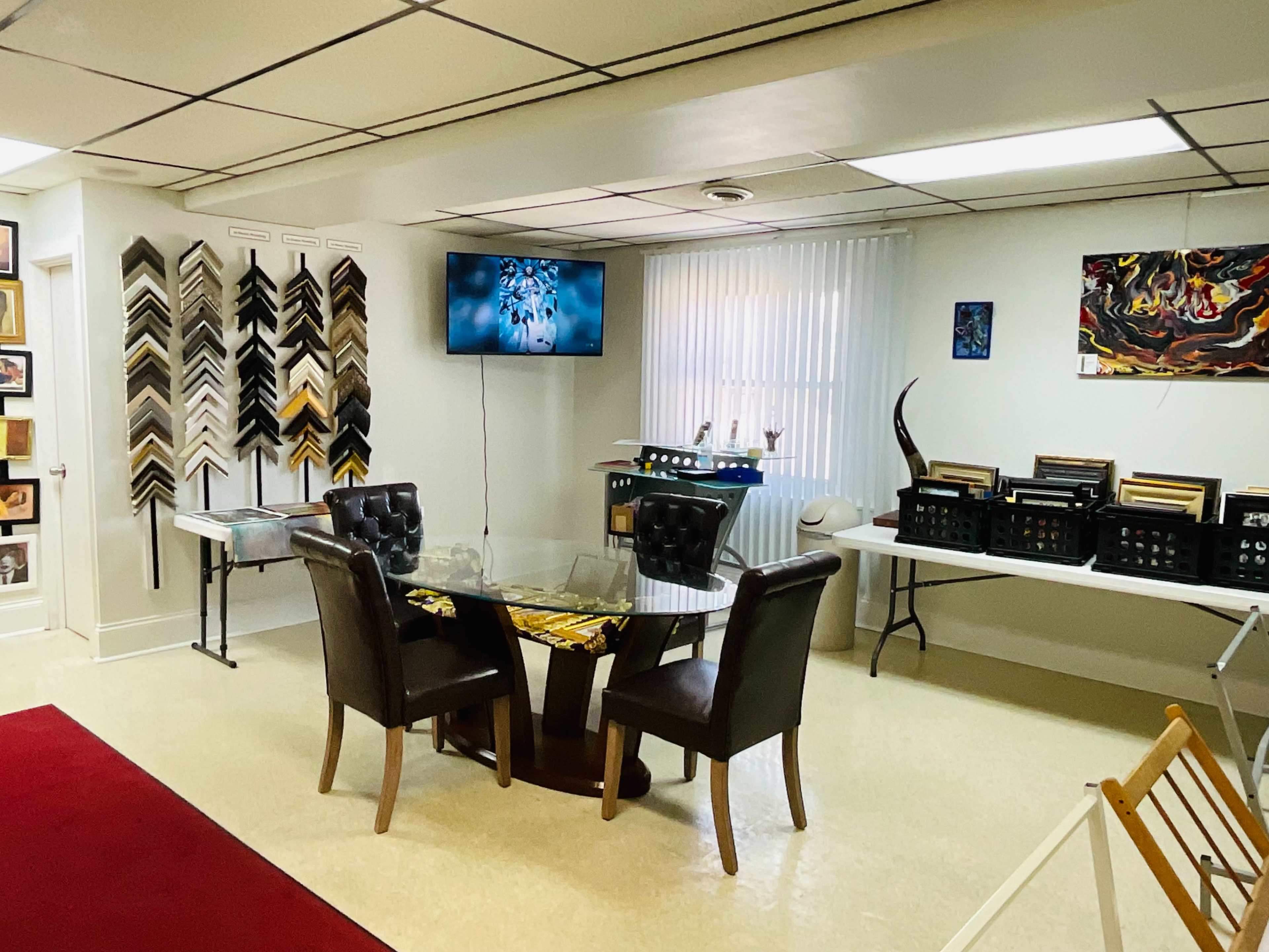 The image shows a room with a glass table and two chairs, a wall of picture frames, a television, and tables displaying framed artwork and other items.