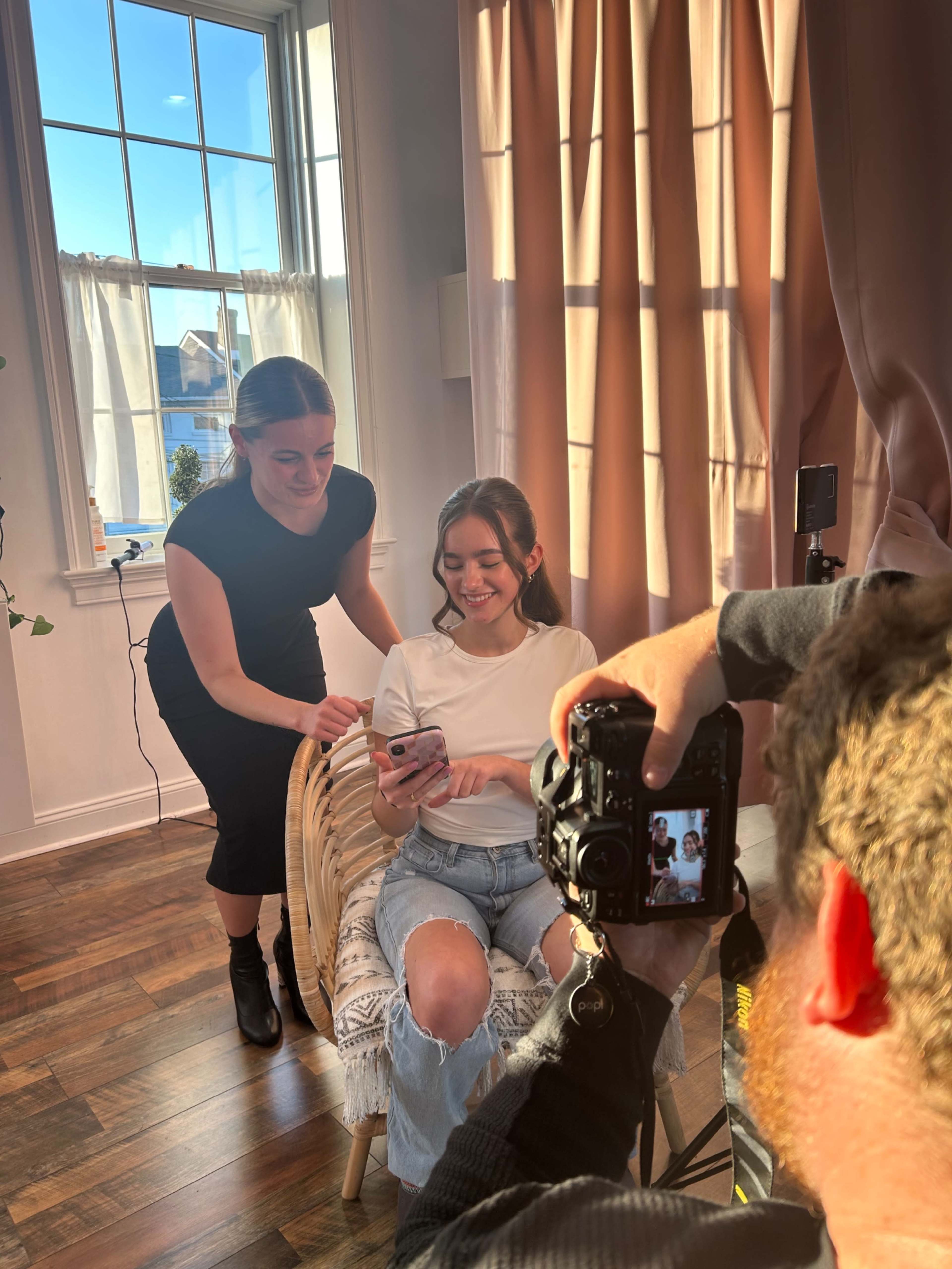 A photographer captures a smiling woman sitting on a chair while another woman helps adjust her hair in a well-lit room.