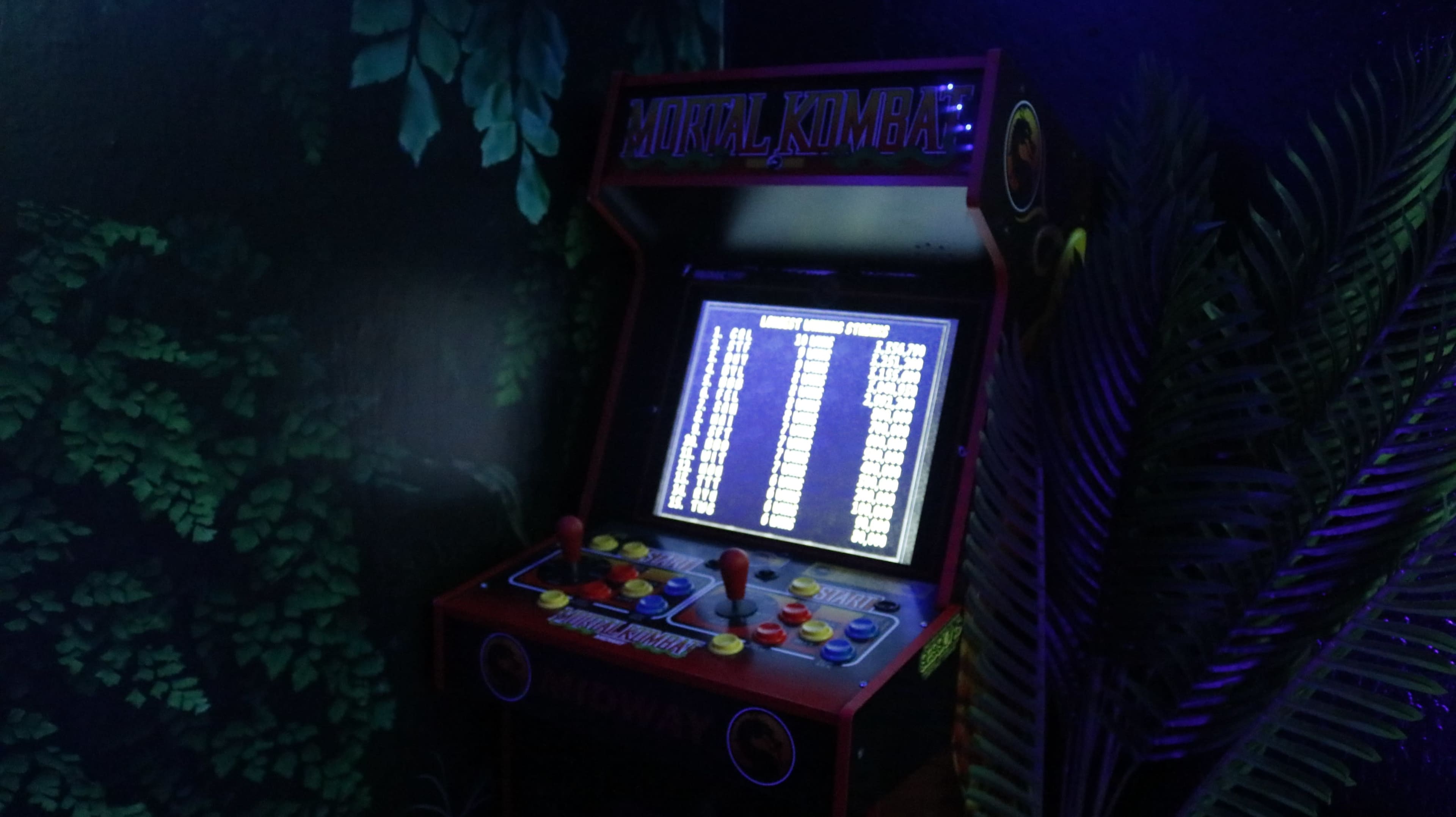 A vintage arcade machine displaying a game menu is situated in a dimly lit room surrounded by leafy decorations.