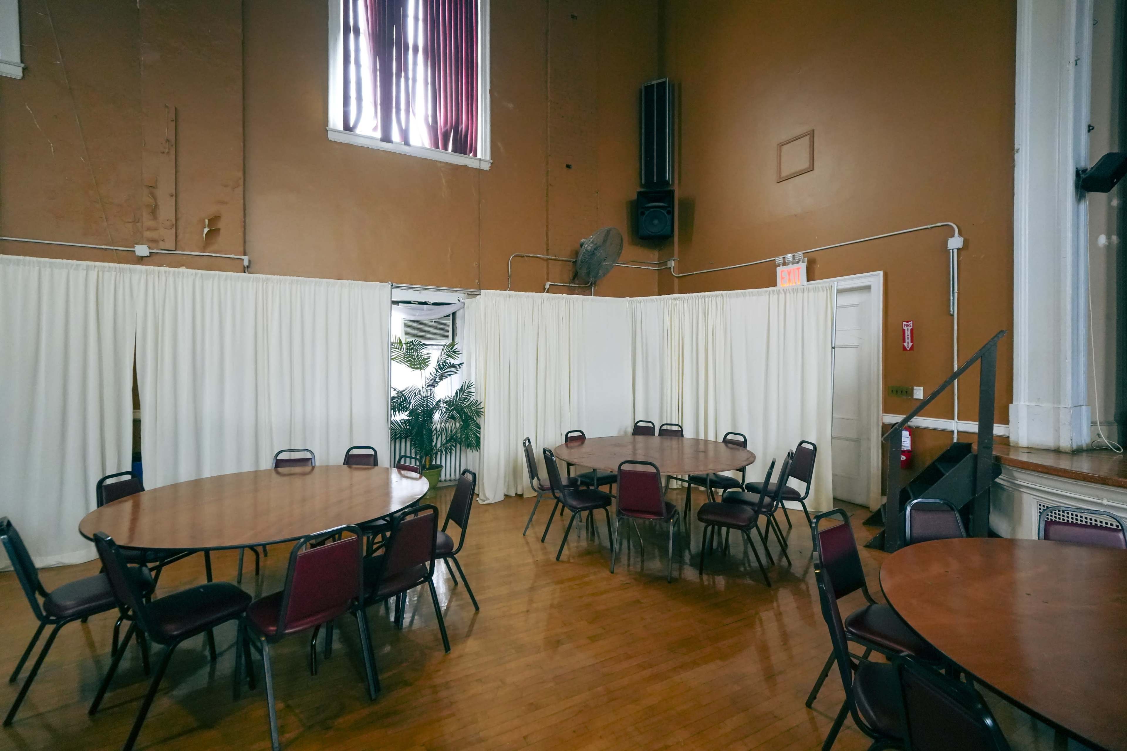 A large, sparsely furnished room with round tables and chairs, featuring a curtain partition and a staircase in the background.