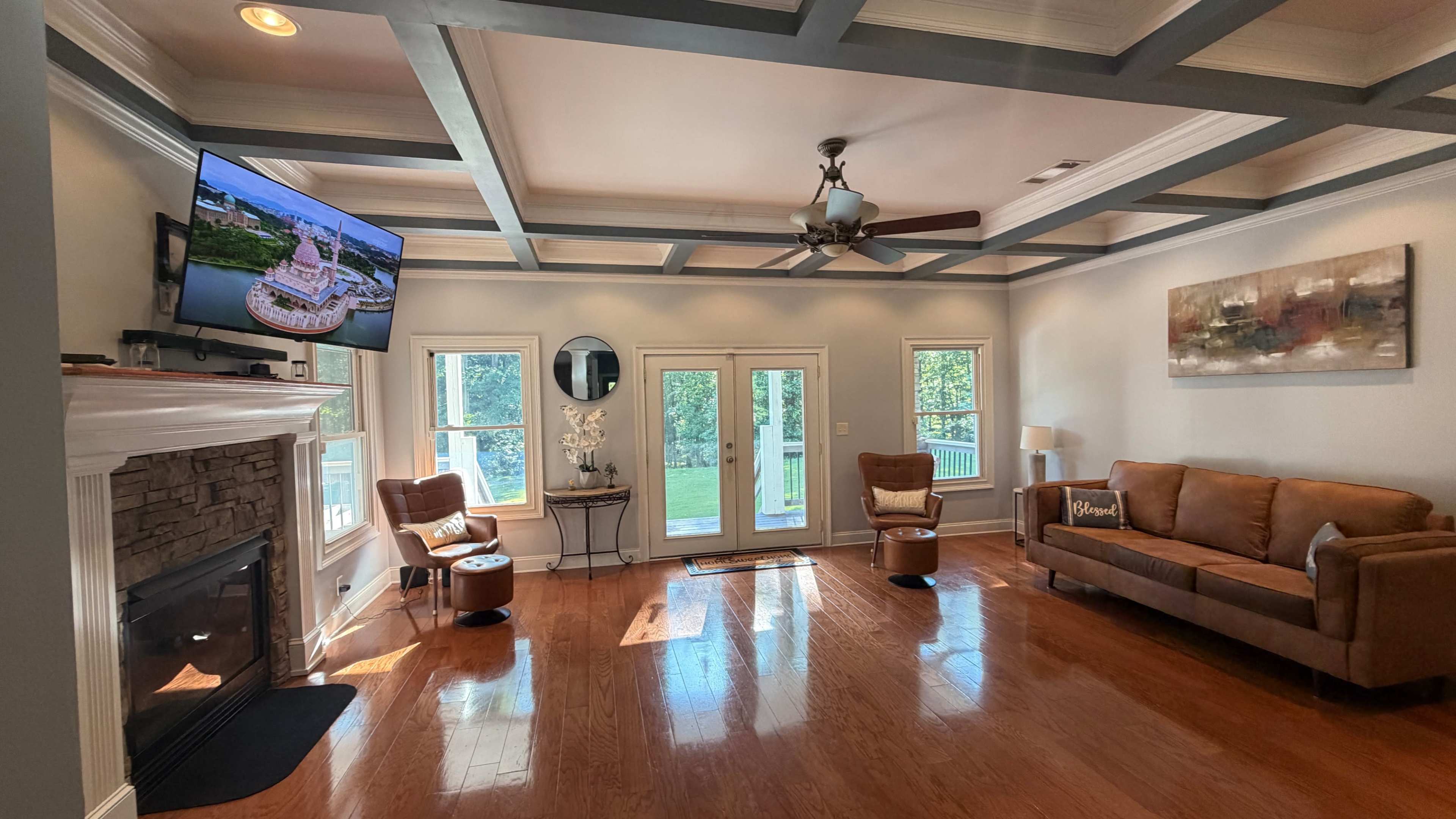 The living room features hardwood floors, a fireplace with stone accents, a brown sofa, two armchairs, and a television mounted on the wall, with large windows providing access to the outdoors.