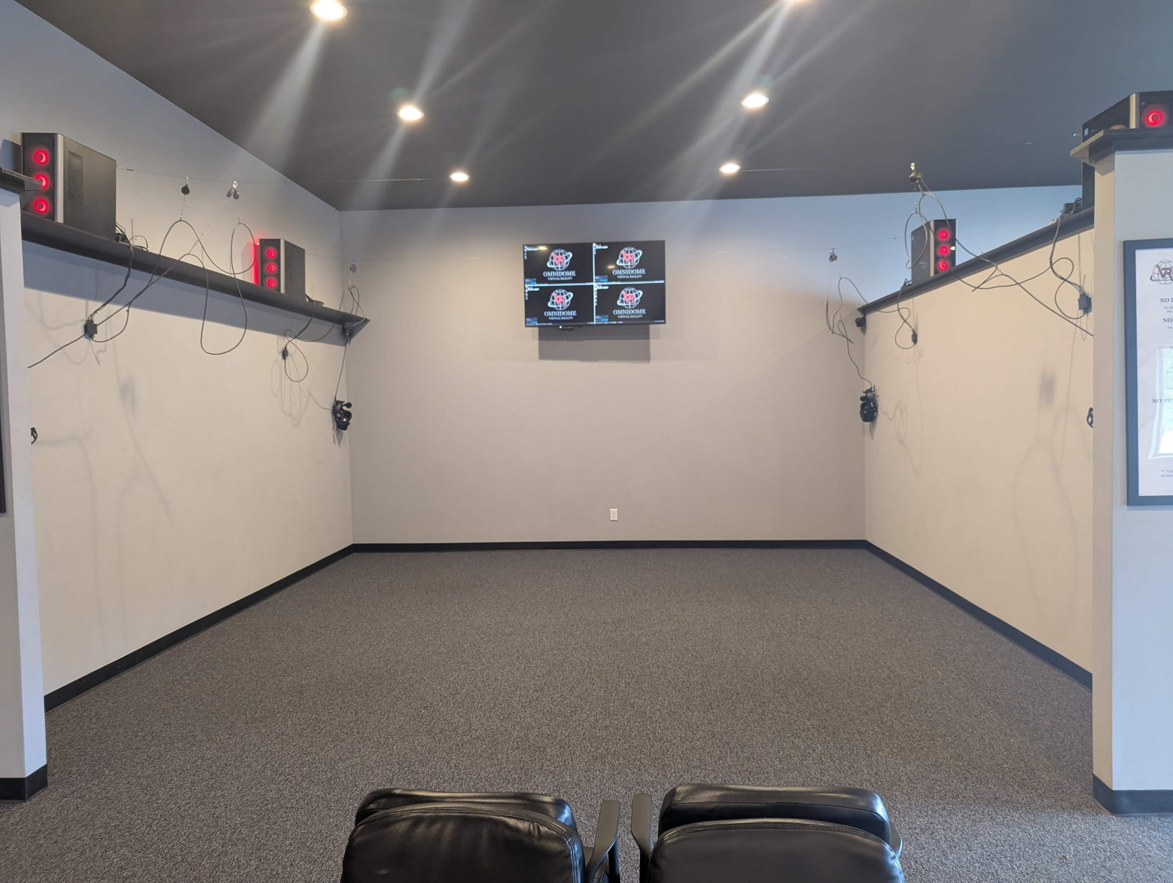 The image shows a spacious room with gray walls, equipped with multiple wall-mounted screens and audio equipment, along with two black chairs positioned in front.