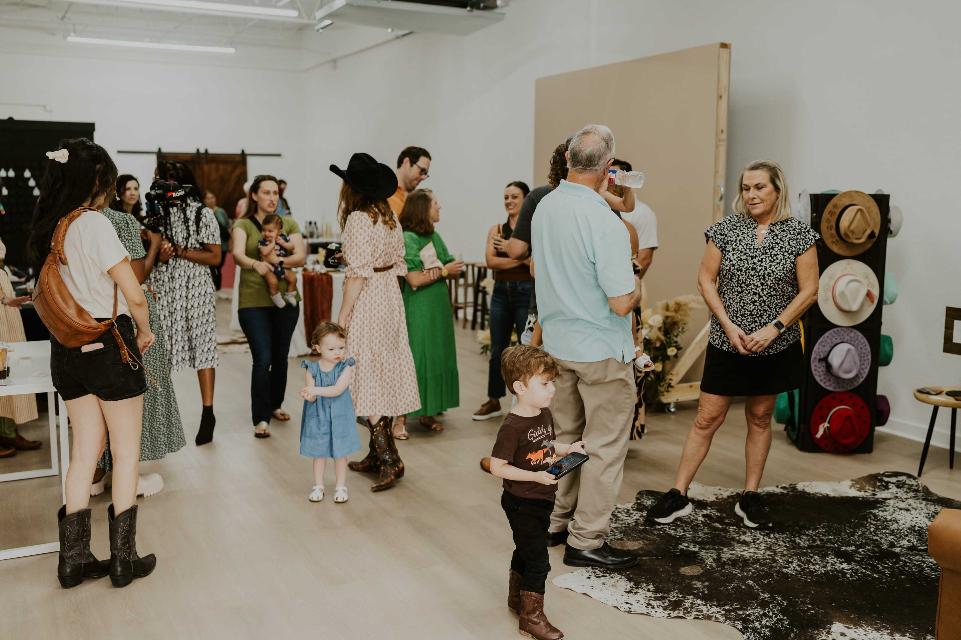 A diverse group of people gathers in a spacious studio, engaging in conversation and examining items displayed on tables, with various hats and decor in the background.