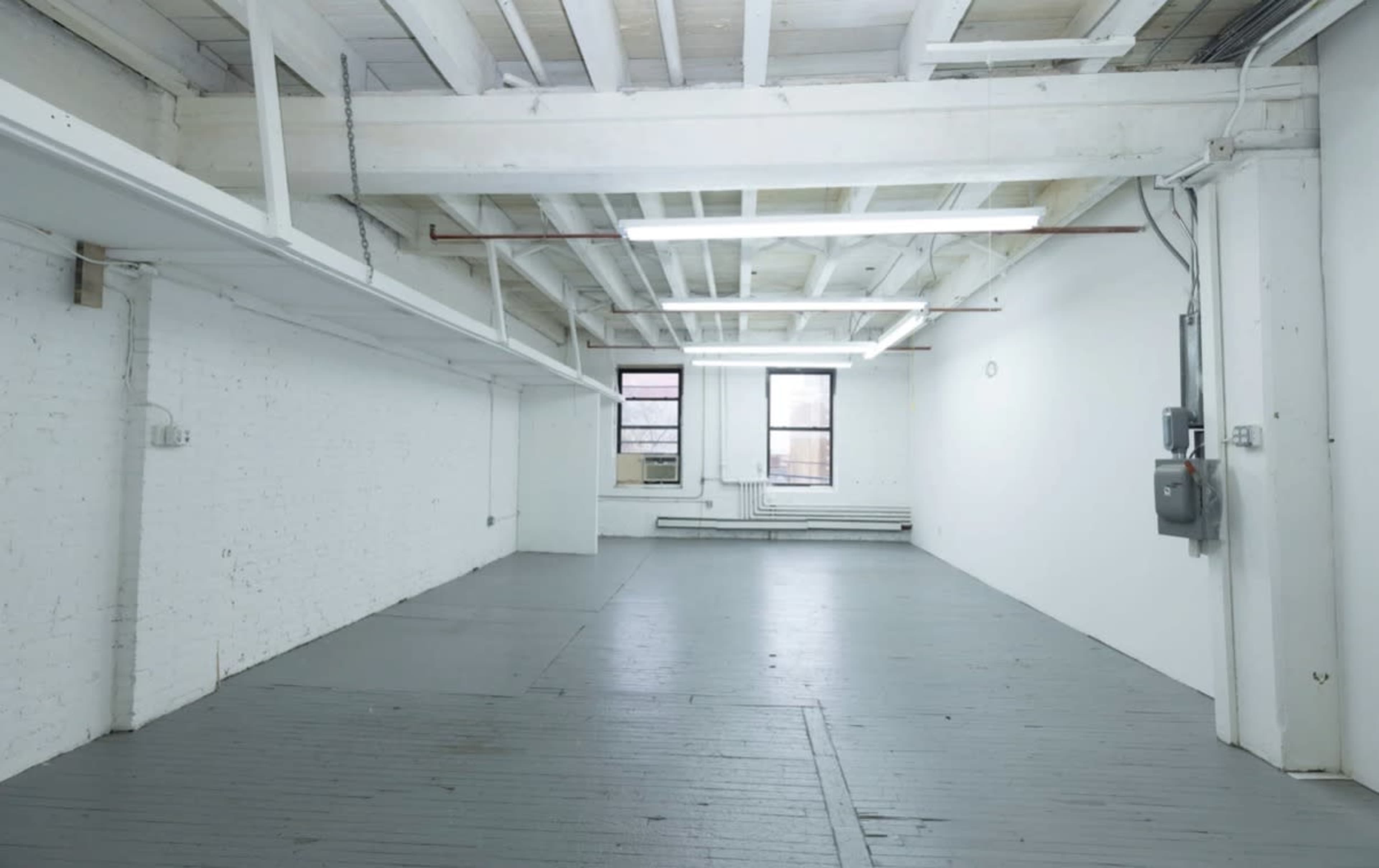 The image shows an empty, spacious room with exposed beams, white walls, and gray flooring, illuminated by fluorescent lights.