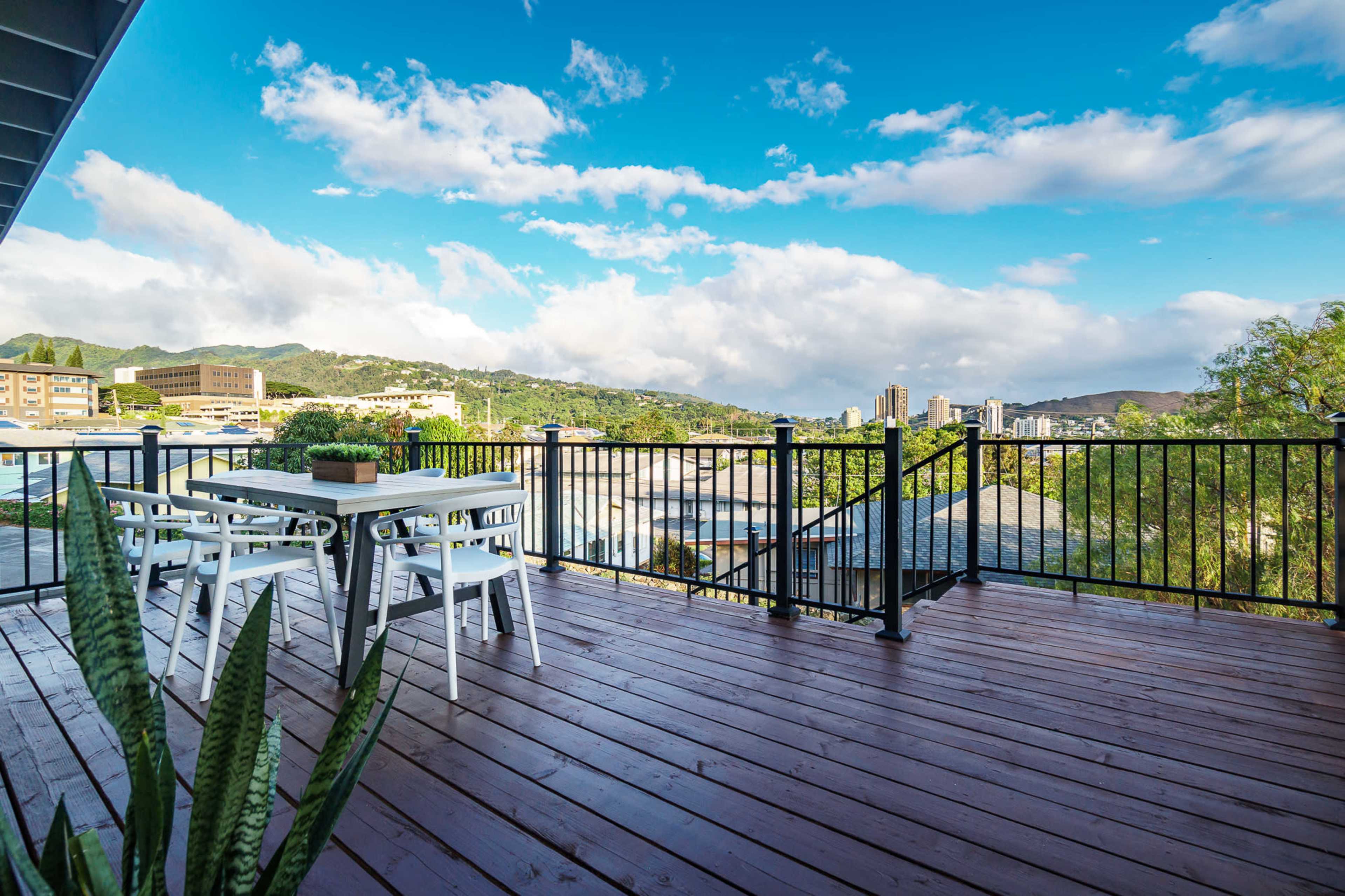 A wooden deck with a dining table overlooks a cityscape and distant hills under a blue sky.