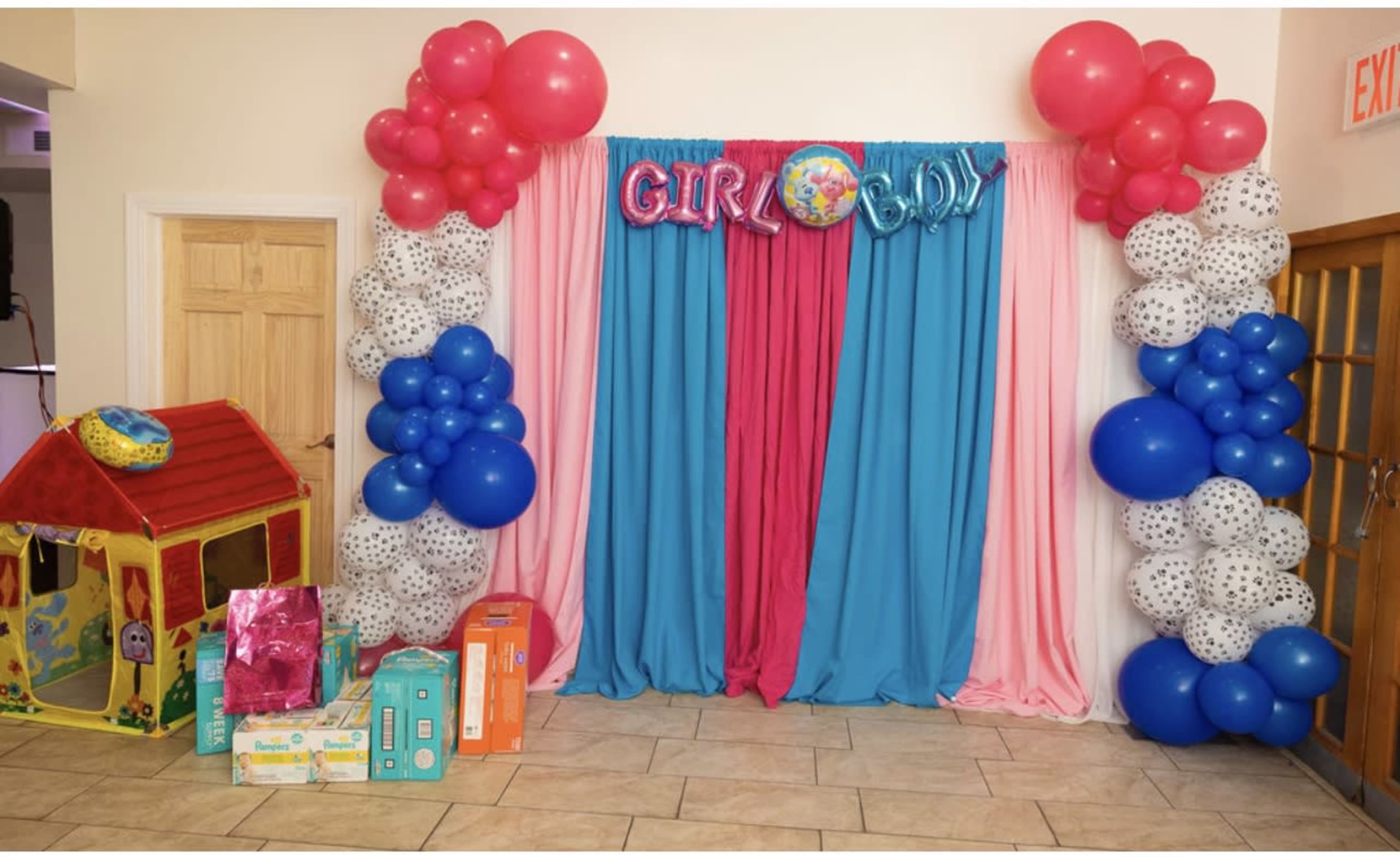 A birthday party setup with a backdrop of pink and blue drapes, balloon decorations on either side, and a small playhouse along with gift boxes arranged in front.