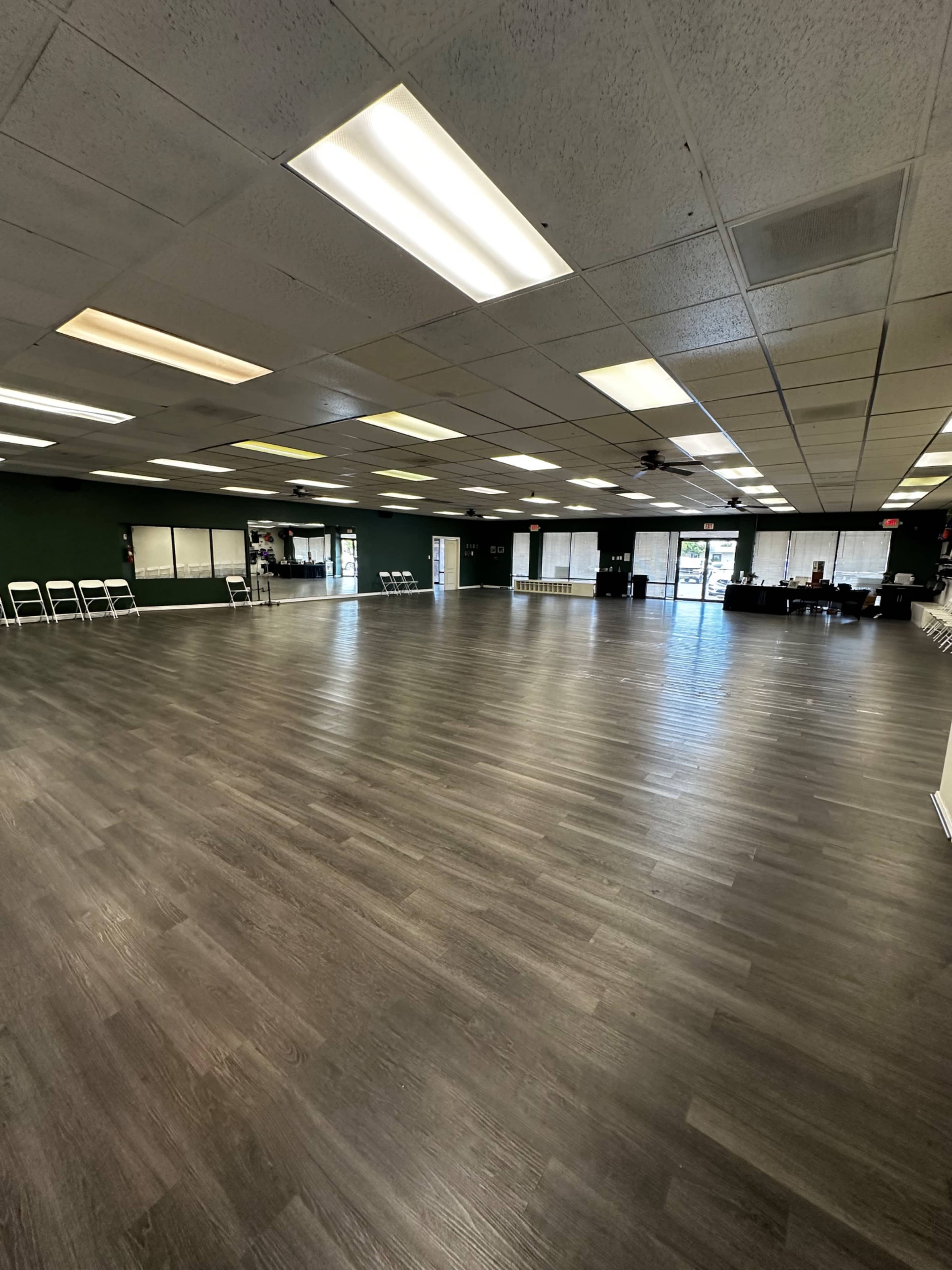 A large, empty room with wooden flooring, bright overhead lights, and several rows of white chairs arranged along the walls.