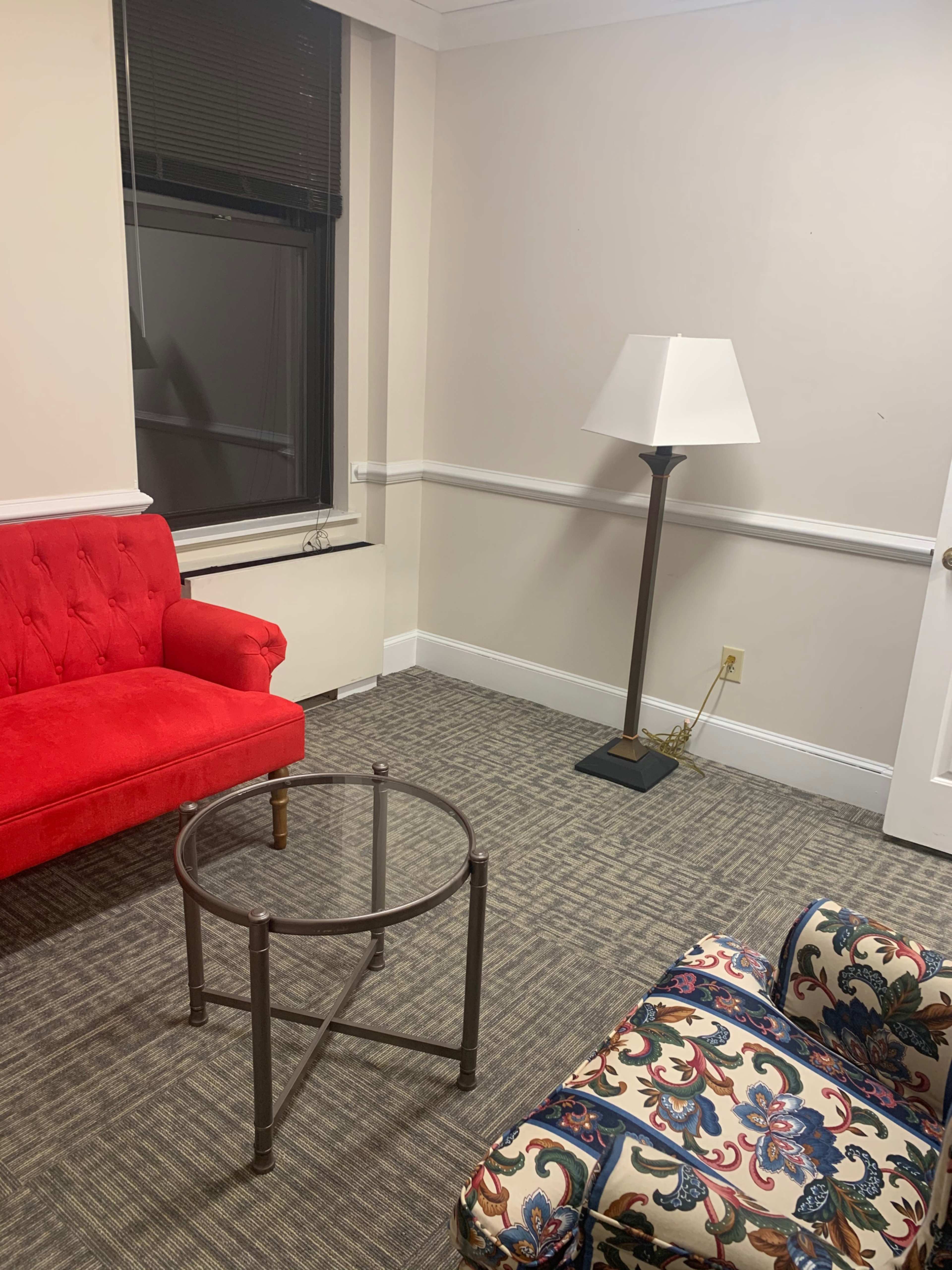 The image shows a small room with a red sofa, a patterned chair, a round glass table, and a floor lamp.