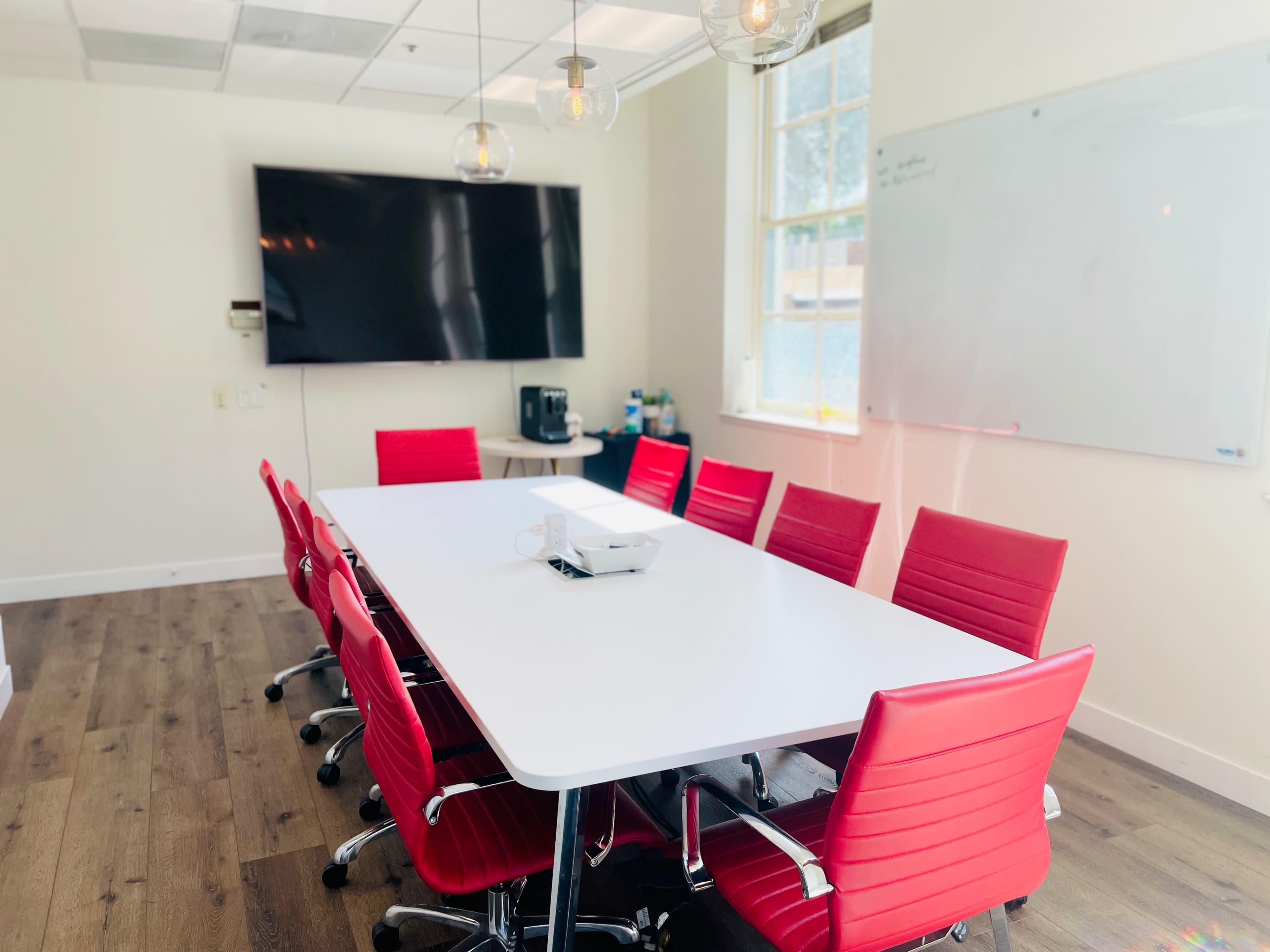 A conference room features a long white table surrounded by red chairs, with a large screen on one wall and a whiteboard on another.