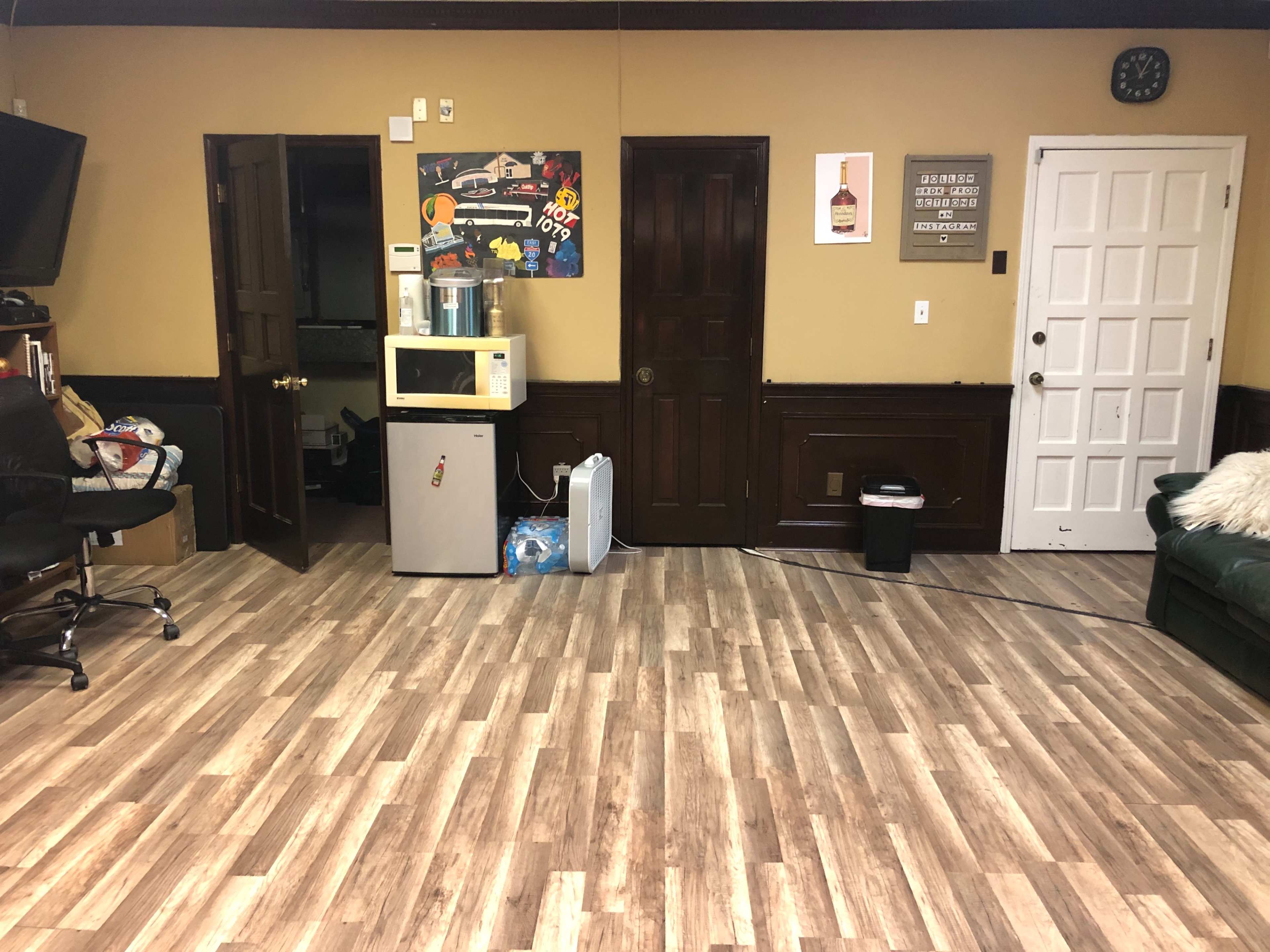 The image shows a room with wooden flooring, featuring a recliner chair, a microwave, a water cooler, and two doors on one wall.