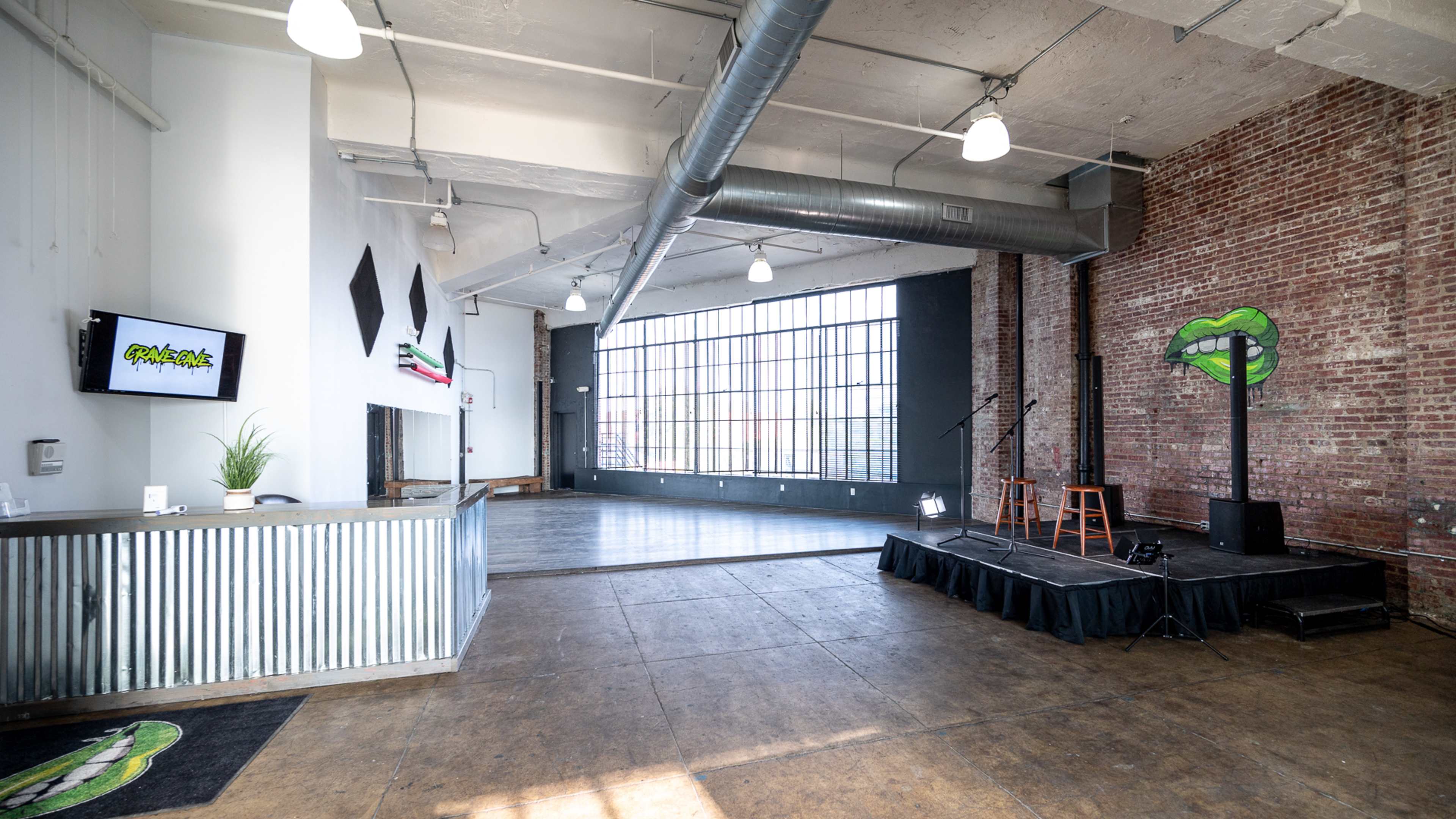 An industrial-style event space with large windows, exposed brick walls, and a small stage with two wooden stools.