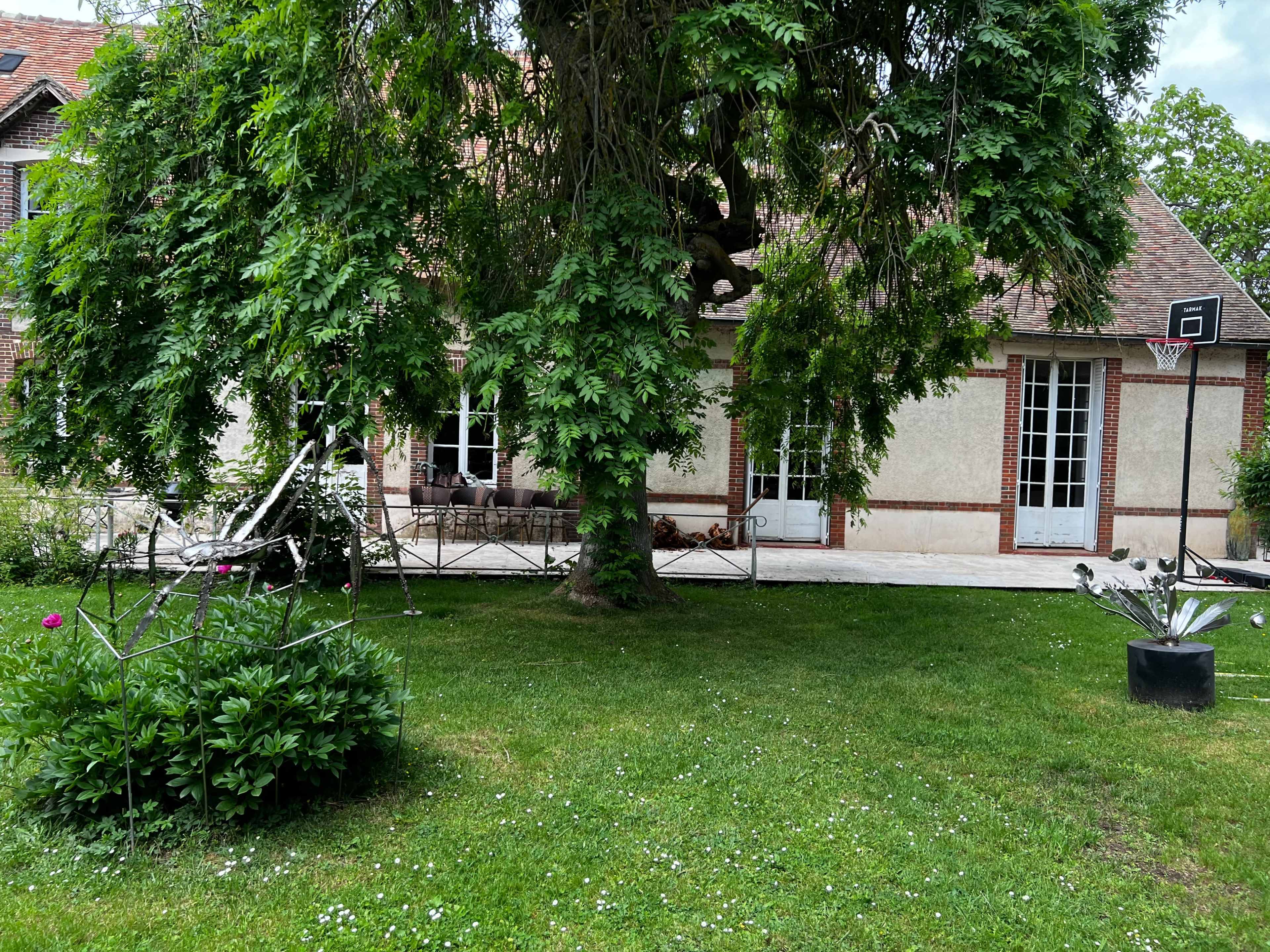 A house with a sloped roof, surrounded by a garden featuring a large tree, a basketball hoop, and decorative plants.