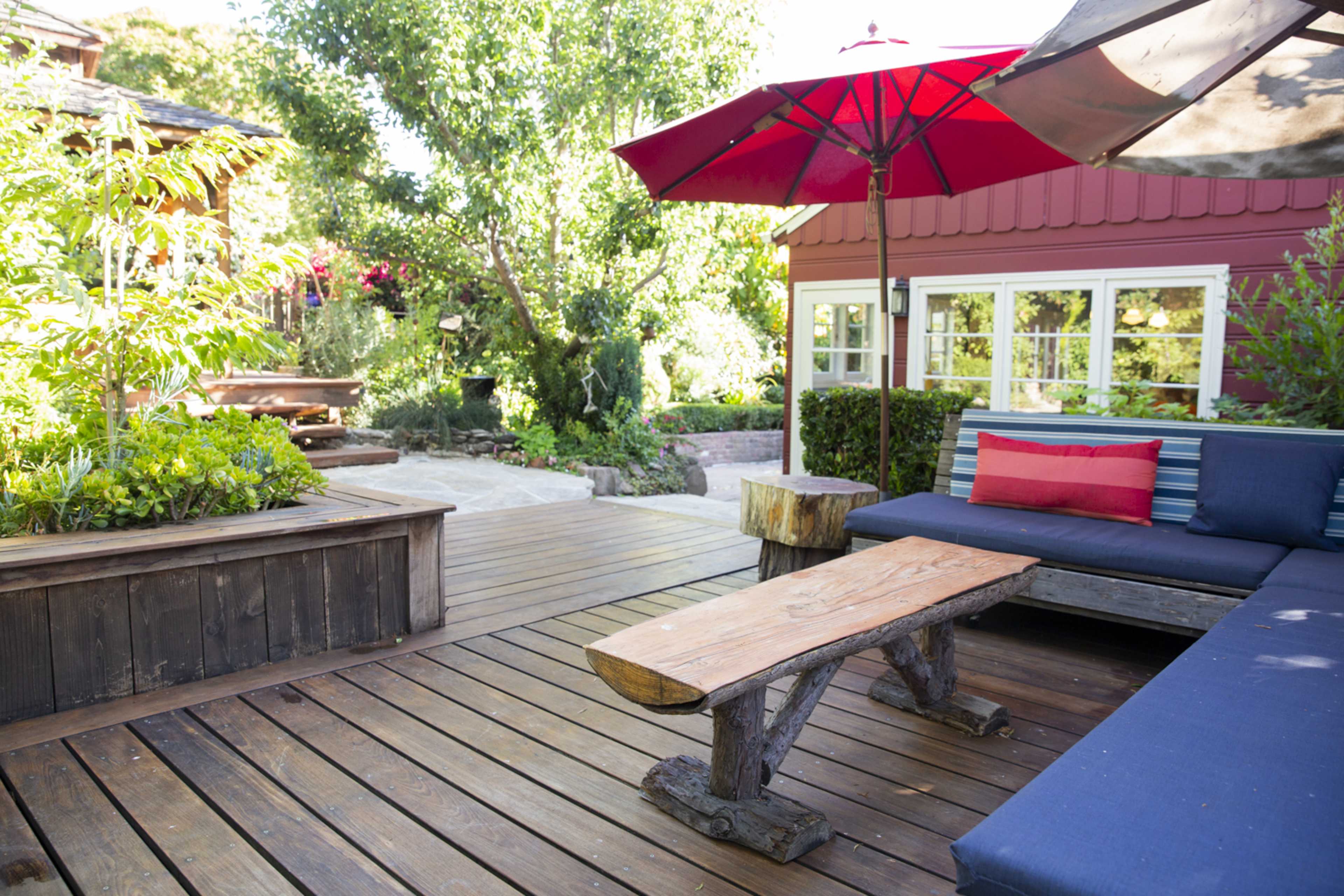 A wooden deck features a seating area with a blue couch and a rustic table beneath a red umbrella, surrounded by greenery and a red building.