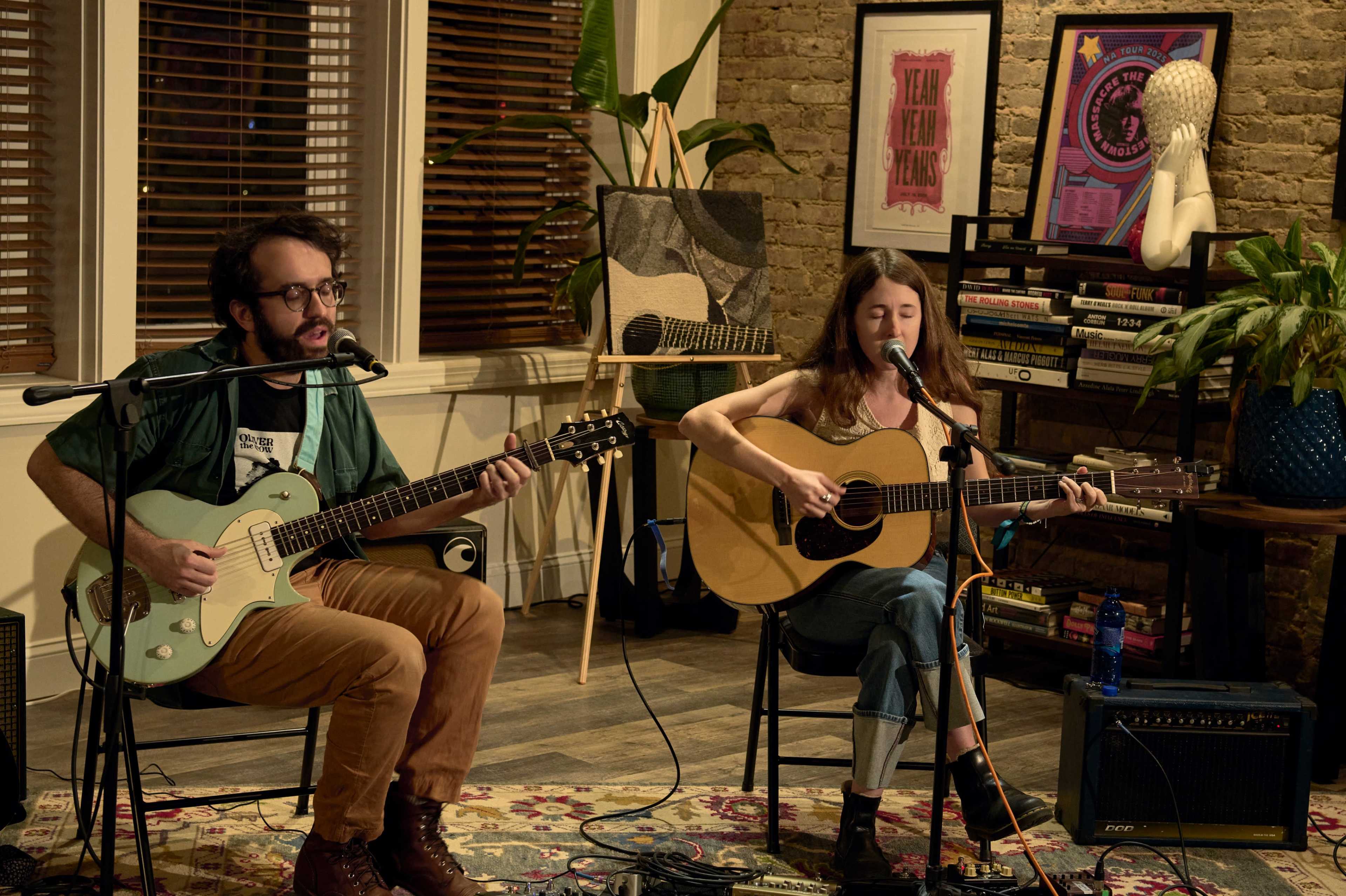 A man and a woman perform music together in a cozy interior setting, with instruments in hand and shelves filled with decor in the background.