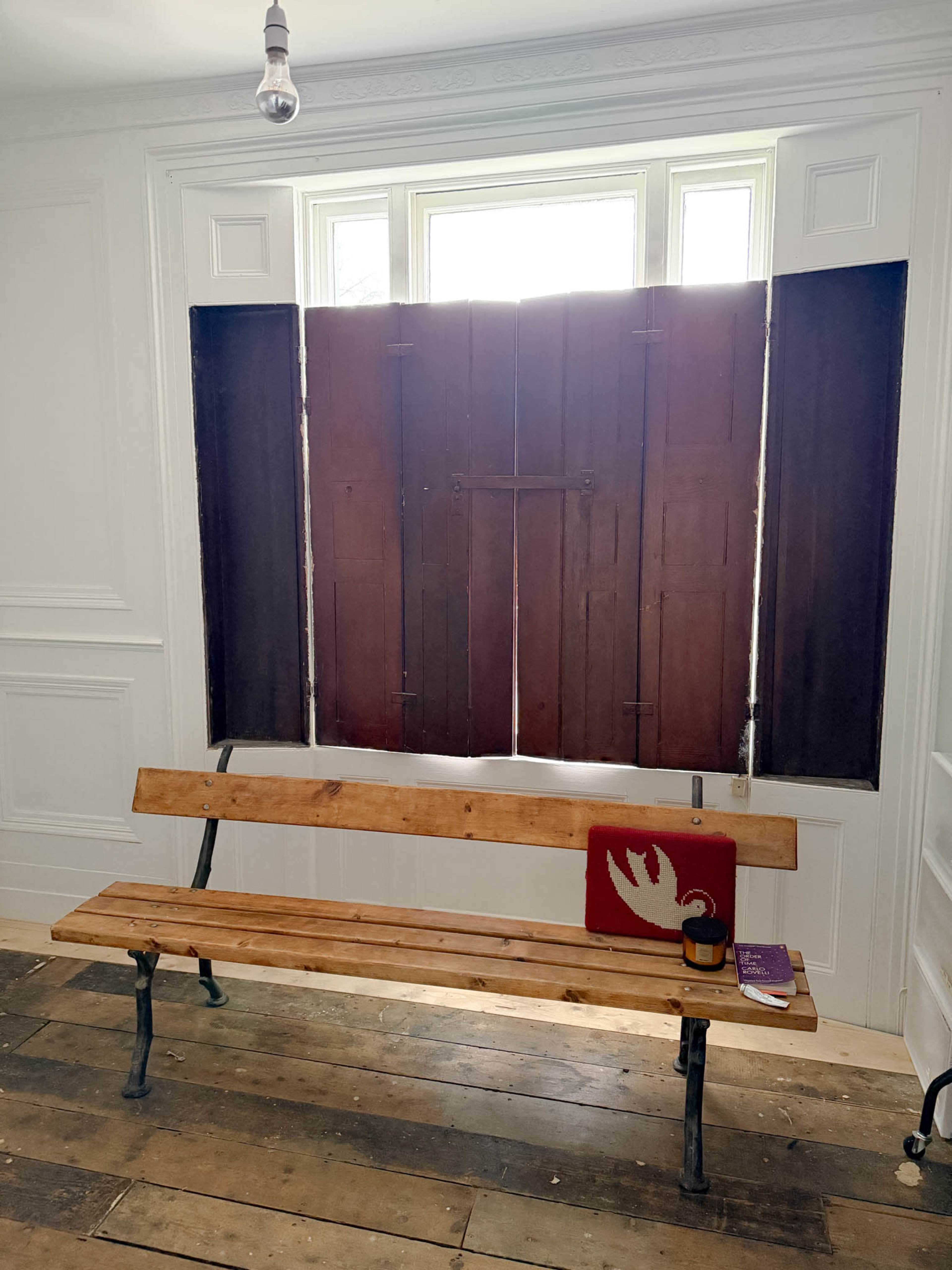 A wooden bench is placed against a wall beside closed wooden shutters, with a small table holding a book and a decorative item.