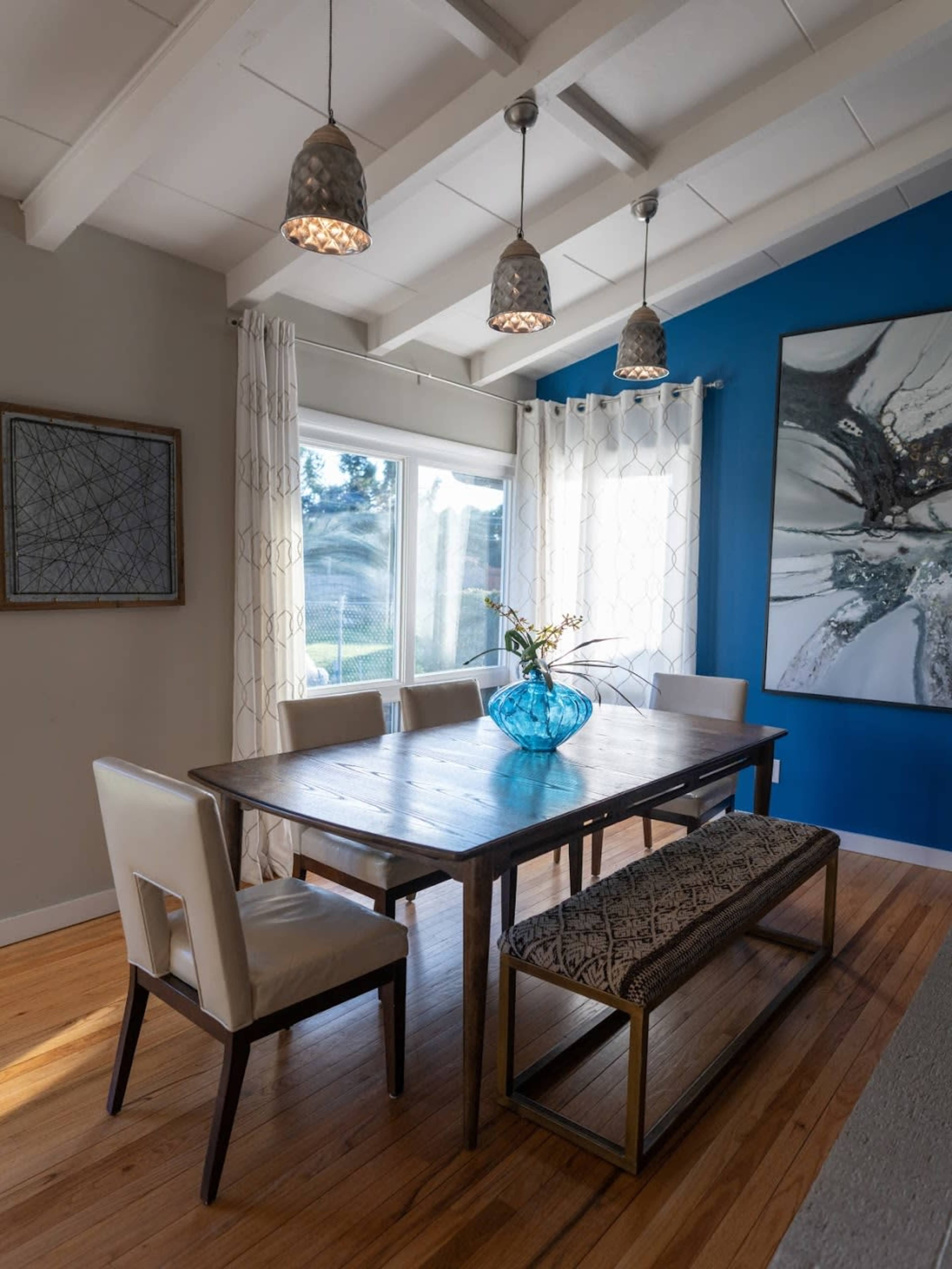 A modern dining room features a wooden table with four chairs and a bench, adorned by decorative light fixtures and wall art.